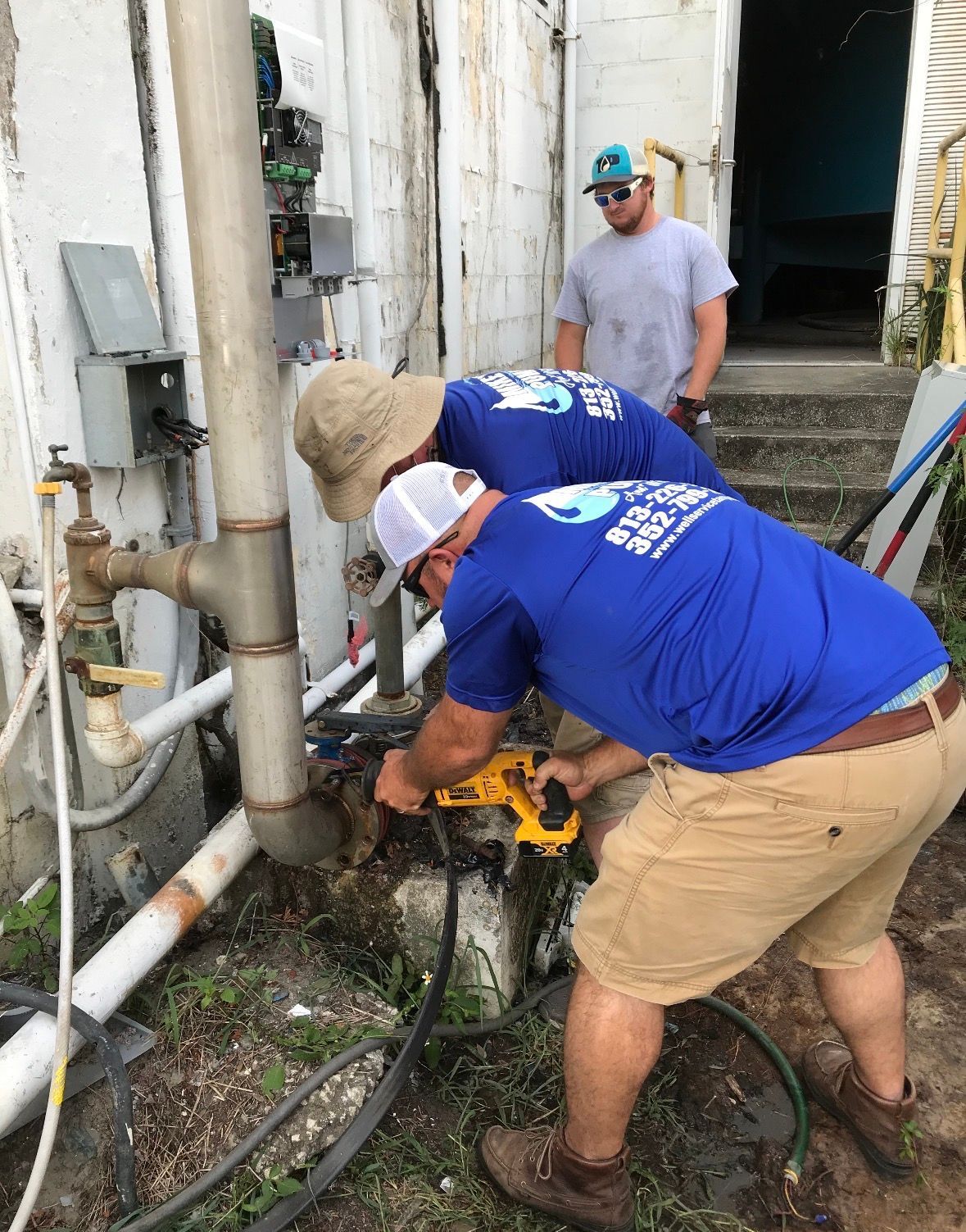 Workers using a tool to cut a pipe beside a building, with one person observing nearby.