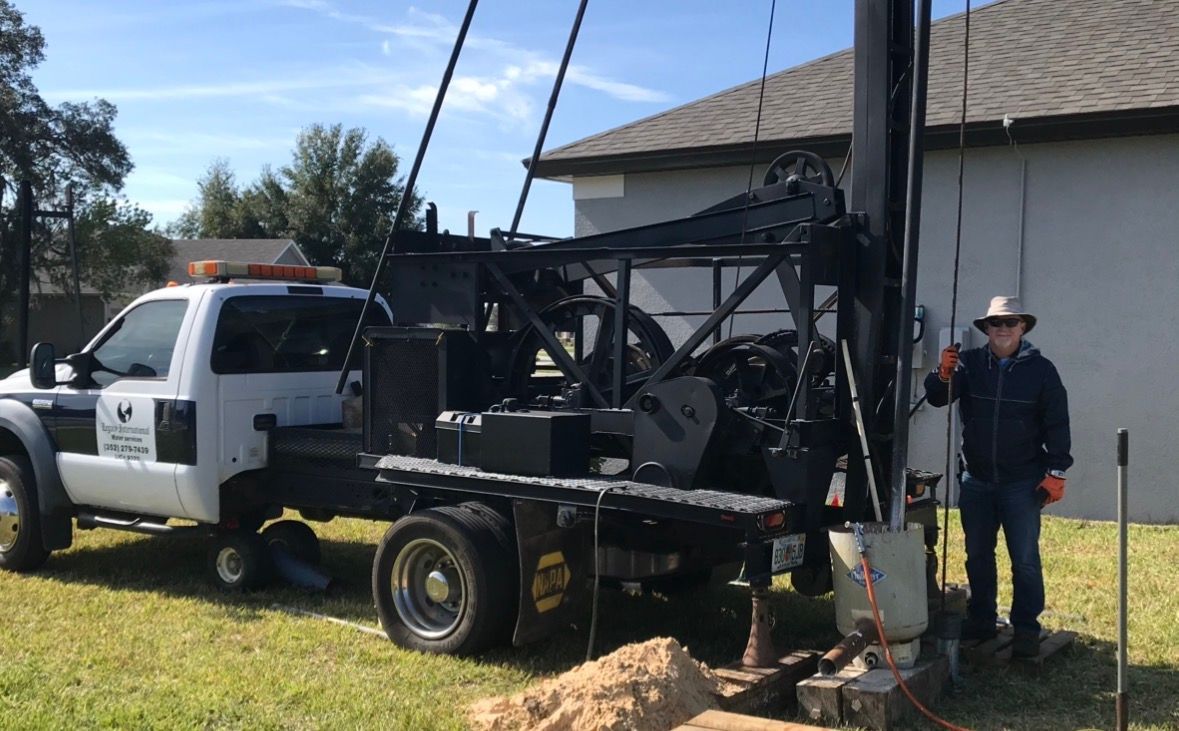 White utility truck with drilling rig parked beside a house; worker stands by the equipment in a grassy yard