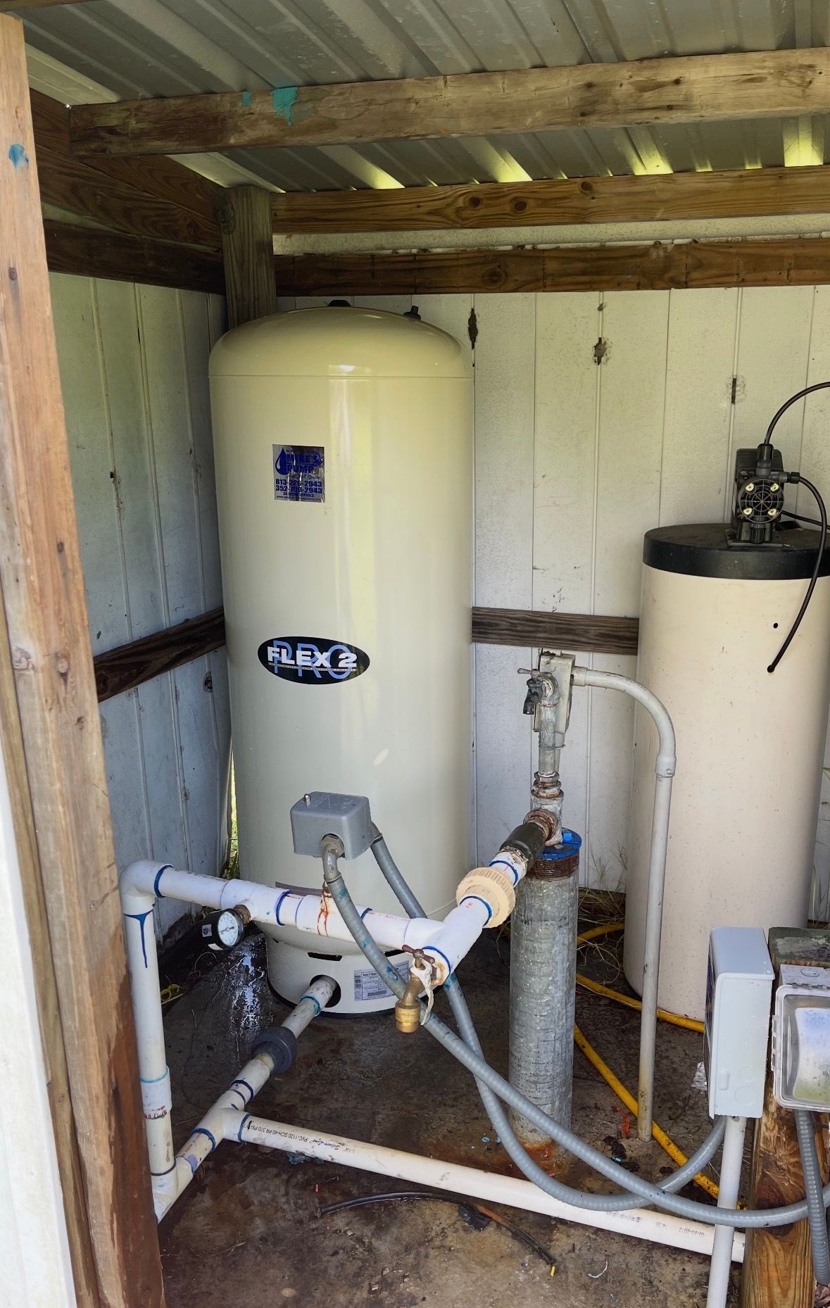 Utility room with water heater, storage tank, and exposed plumbing under a wooden shelter