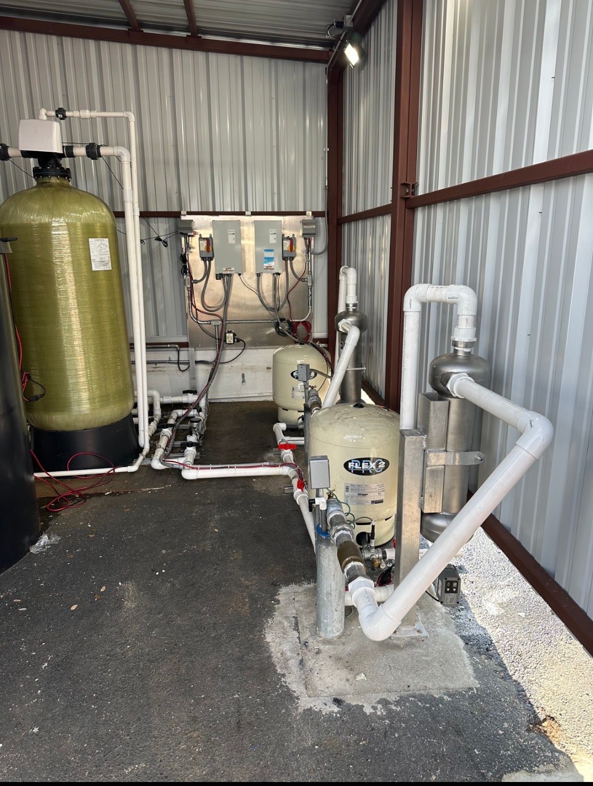 Industrial water treatment equipment in a corrugated metal shed, with tanks, pumps, and white piping on concrete floor