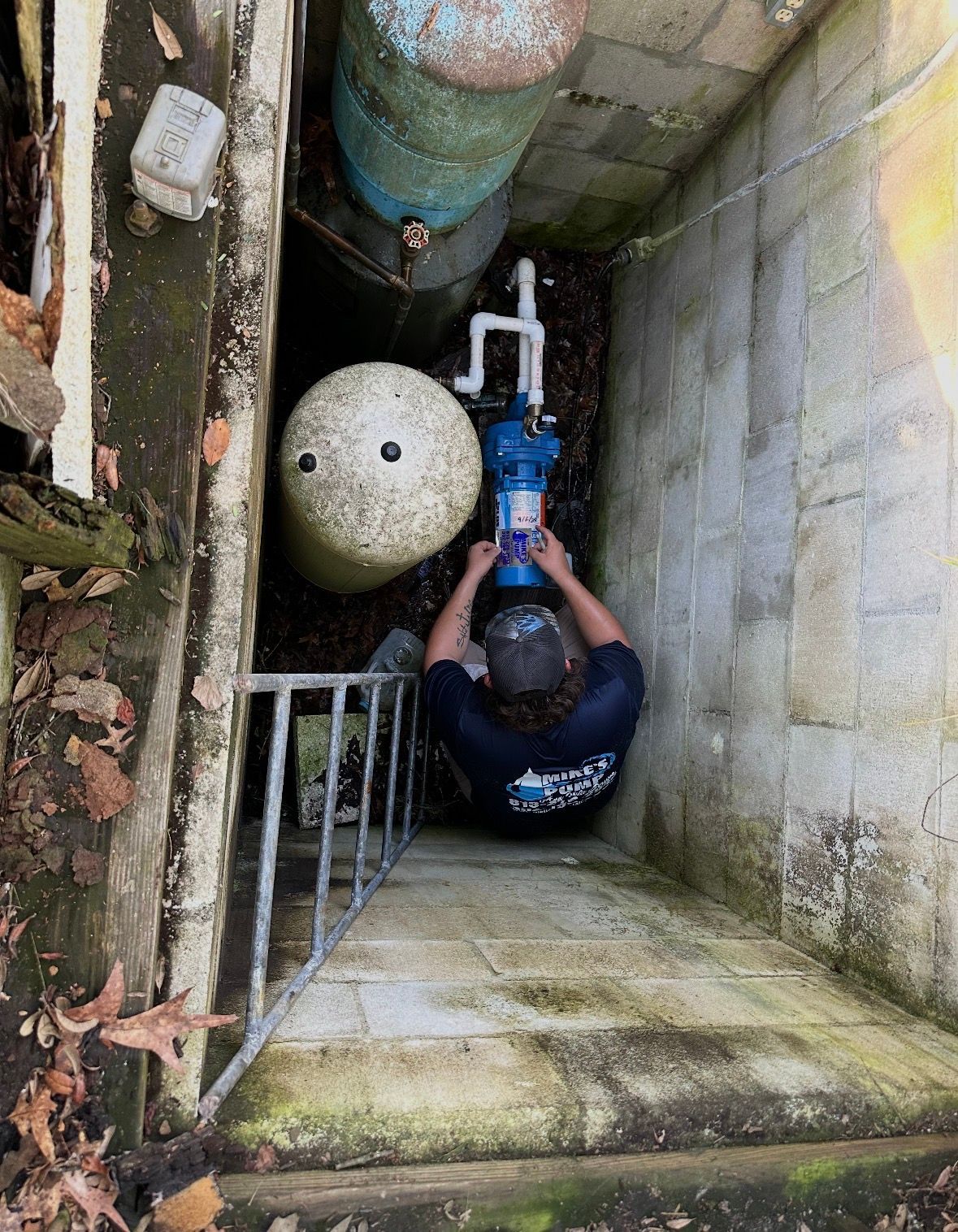 Worker in a narrow utility pit using a blue power tool on pipes and concrete walls.