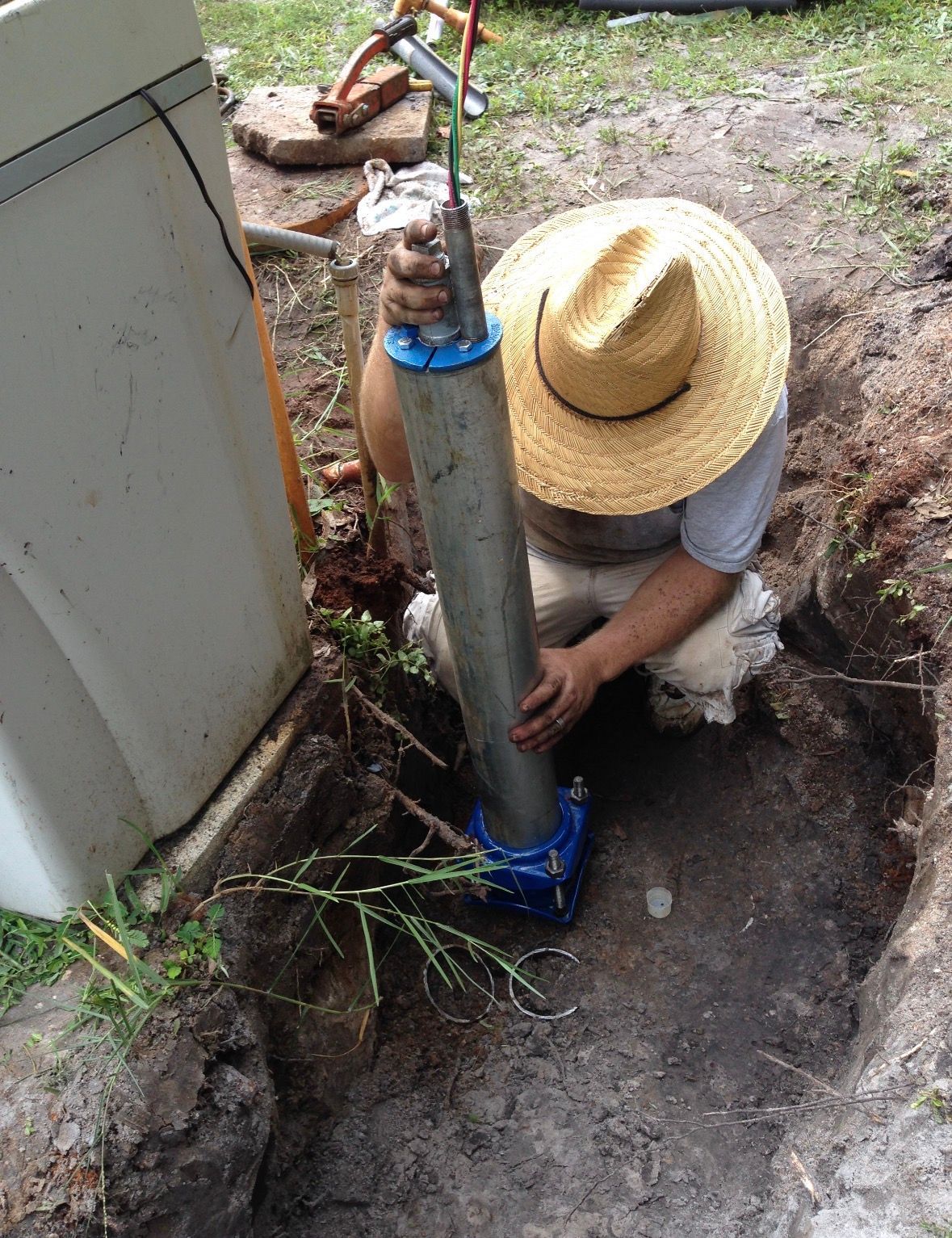 Person in straw hat installing a gray metal post beside a building in a dirt trench