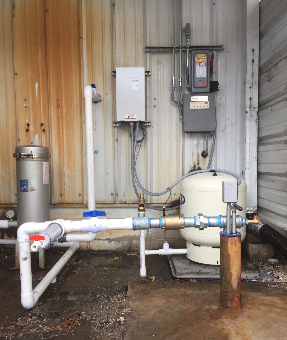 Outdoor industrial plumbing and pump equipment beside a building wall, with pipes, valves, and tanks.