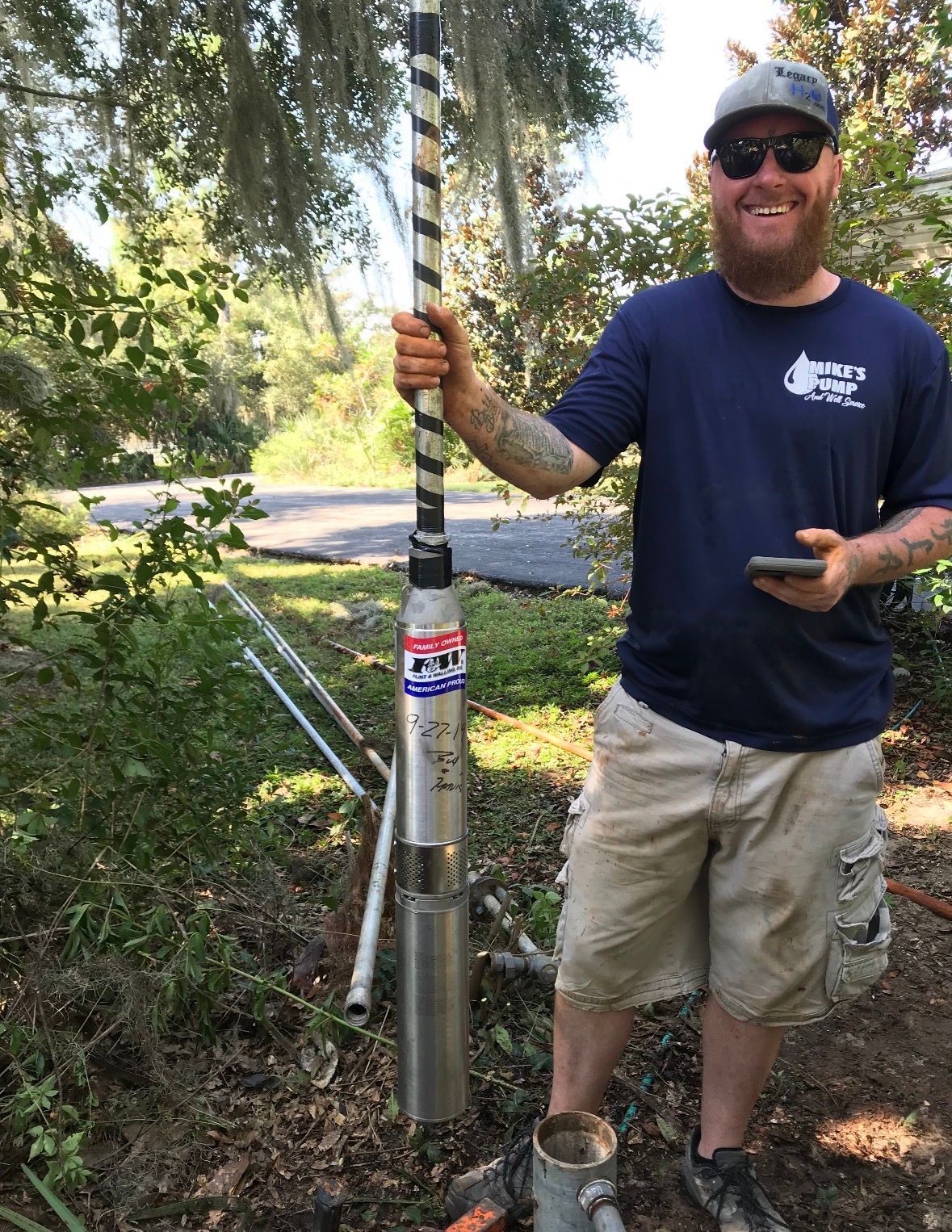 Smiling man in sunglasses holds a tall pole in a wooded yard.