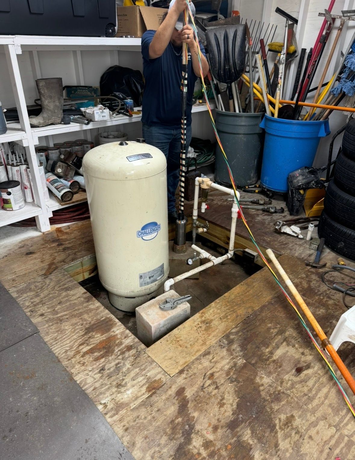 Person using a long-handled tool beside a beige tank in a cluttered workshop, with brooms and buckets nearby