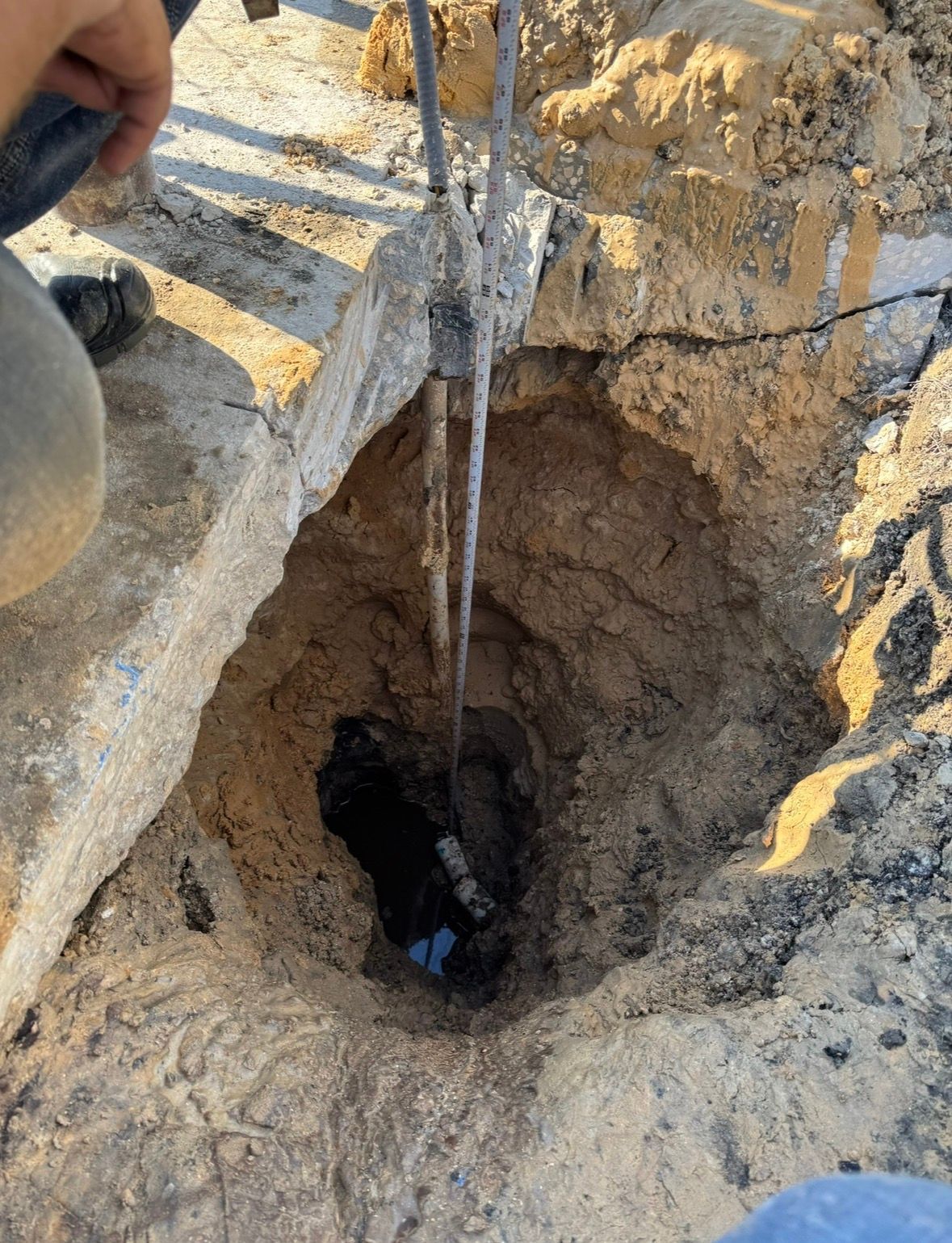 A rope hangs into a deep, narrow hole in rocky ground near a person’s hand.