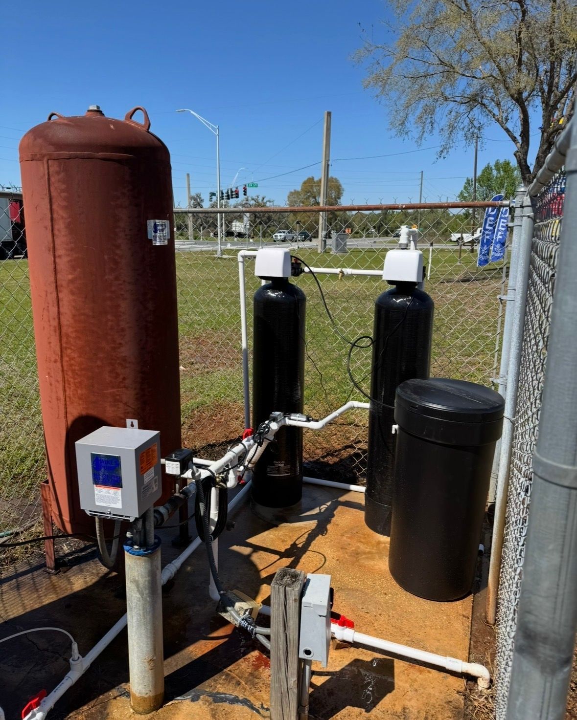 Outdoor water treatment equipment with red and black tanks, pipes, and control box beside a fence