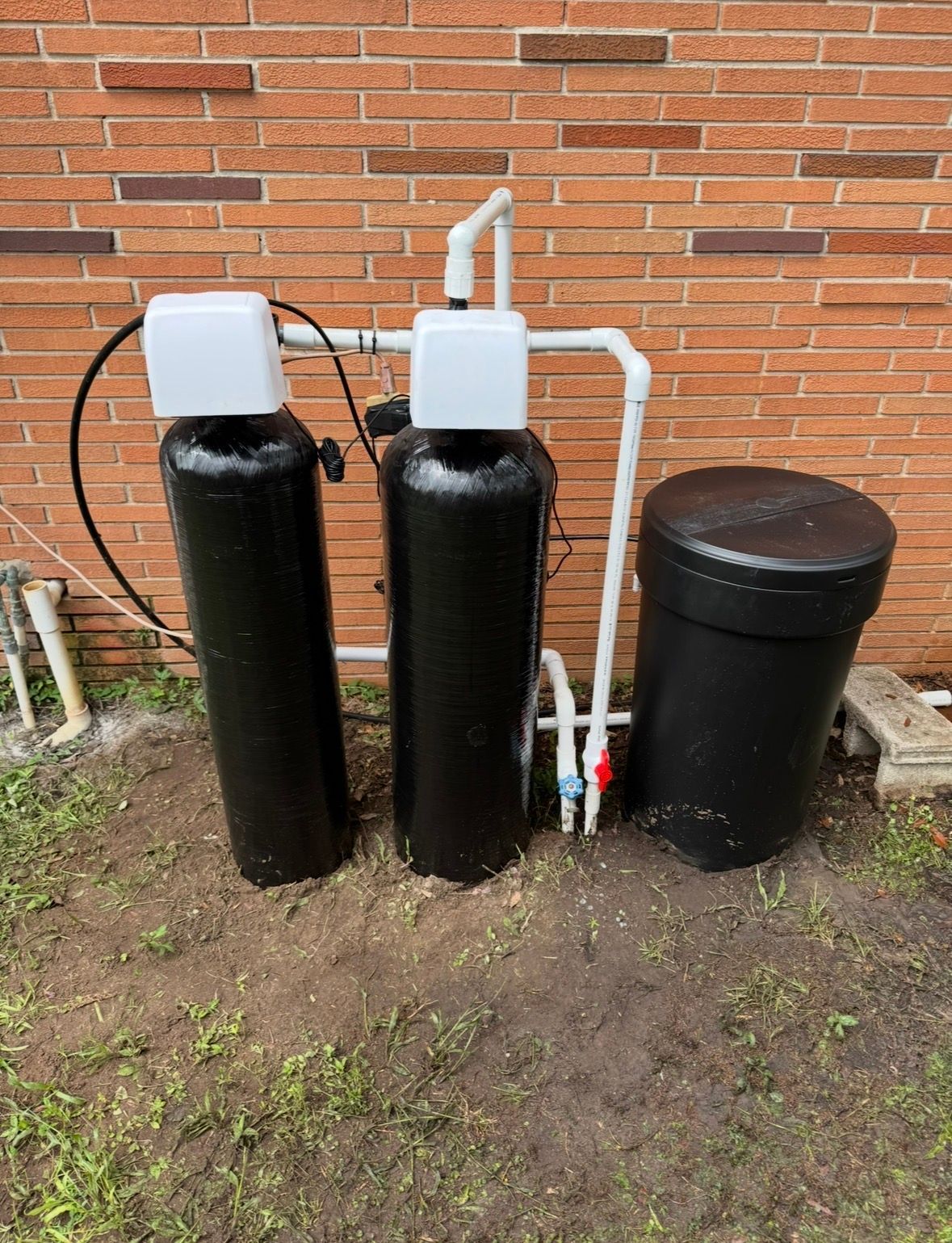 Two black water treatment tanks with white pipes and covers beside a brick wall outdoors.