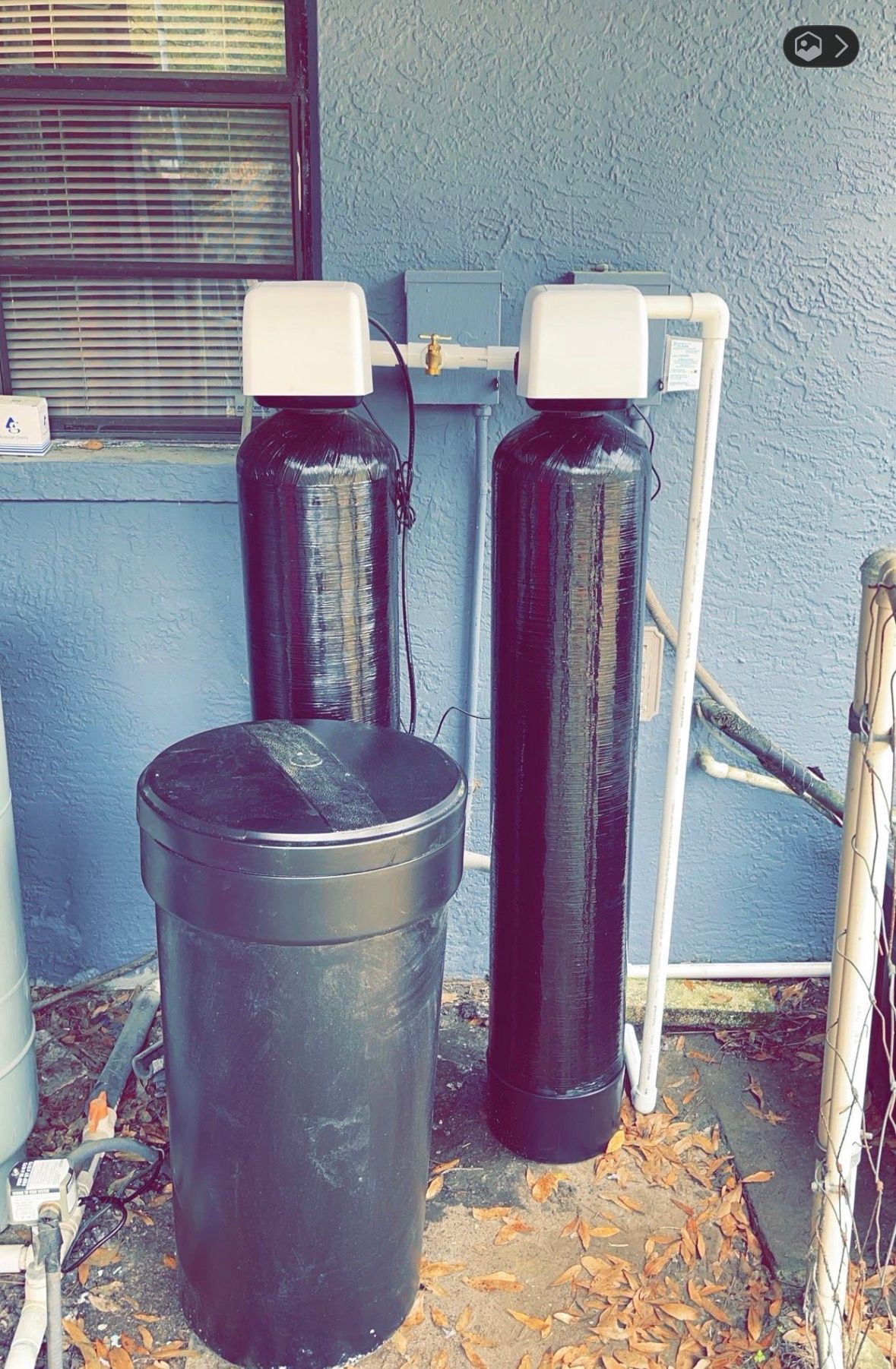 Two large dark water softener tanks beside a trash can against a blue wall outdoors
