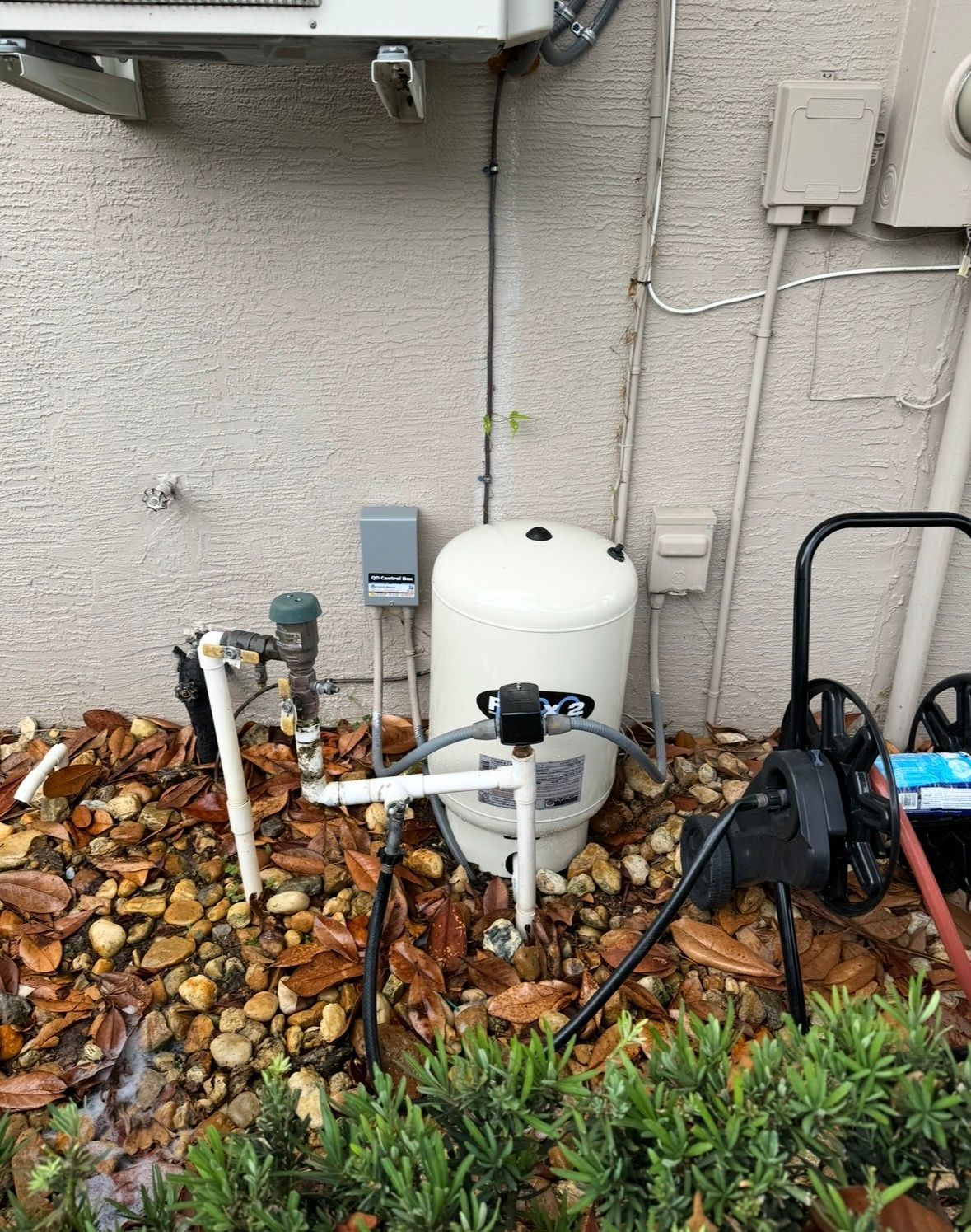 White tank and utility pipes beside a house wall, with fallen leaves and garden plants in front.