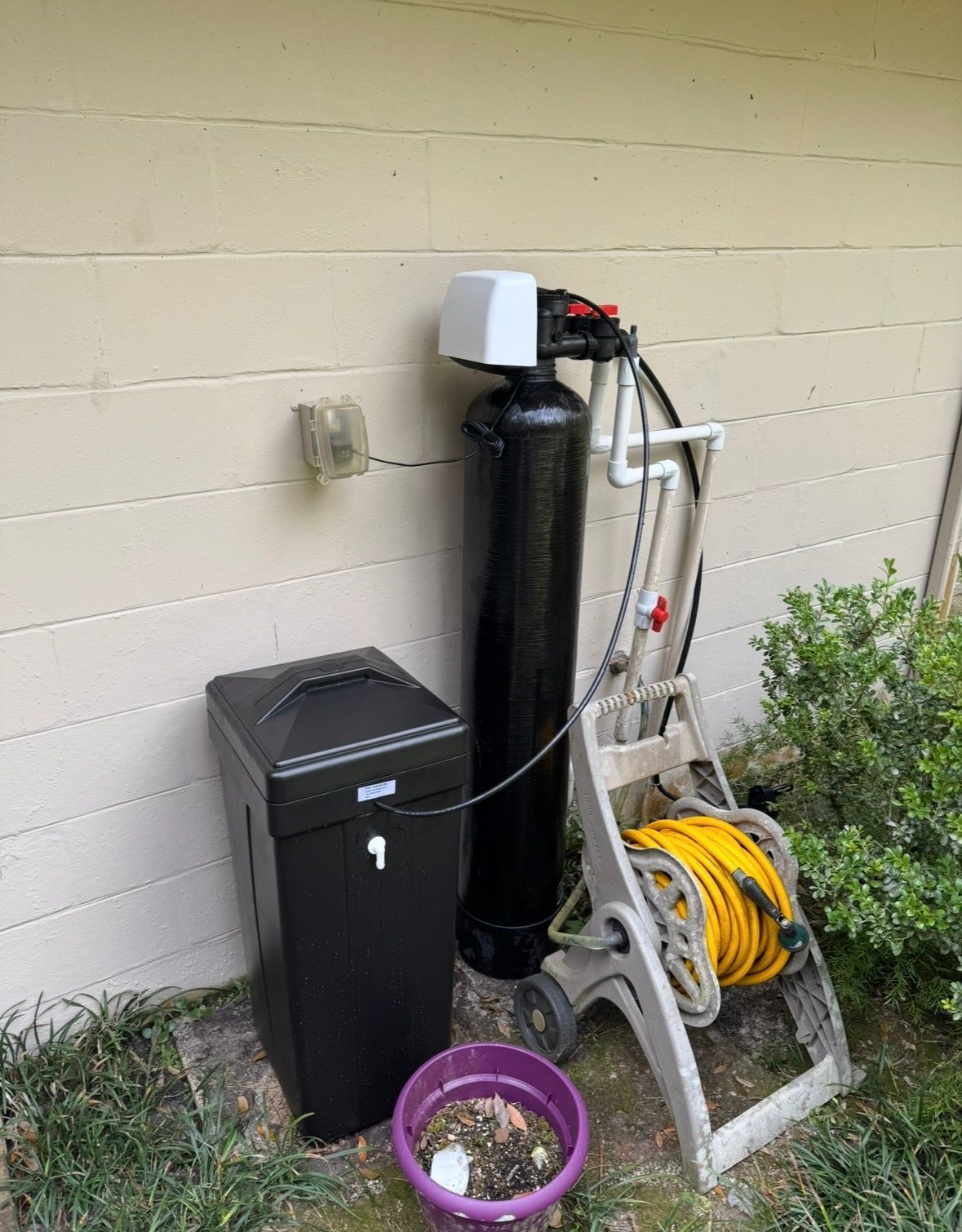 Outdoor equipment beside a beige wall: black bin, water softener tank, hoses, and a ladder with extension cords.
