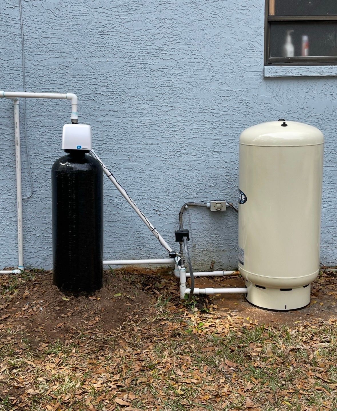 Outdoor utility equipment with a black tank and cream-colored pressure tank beside a house.