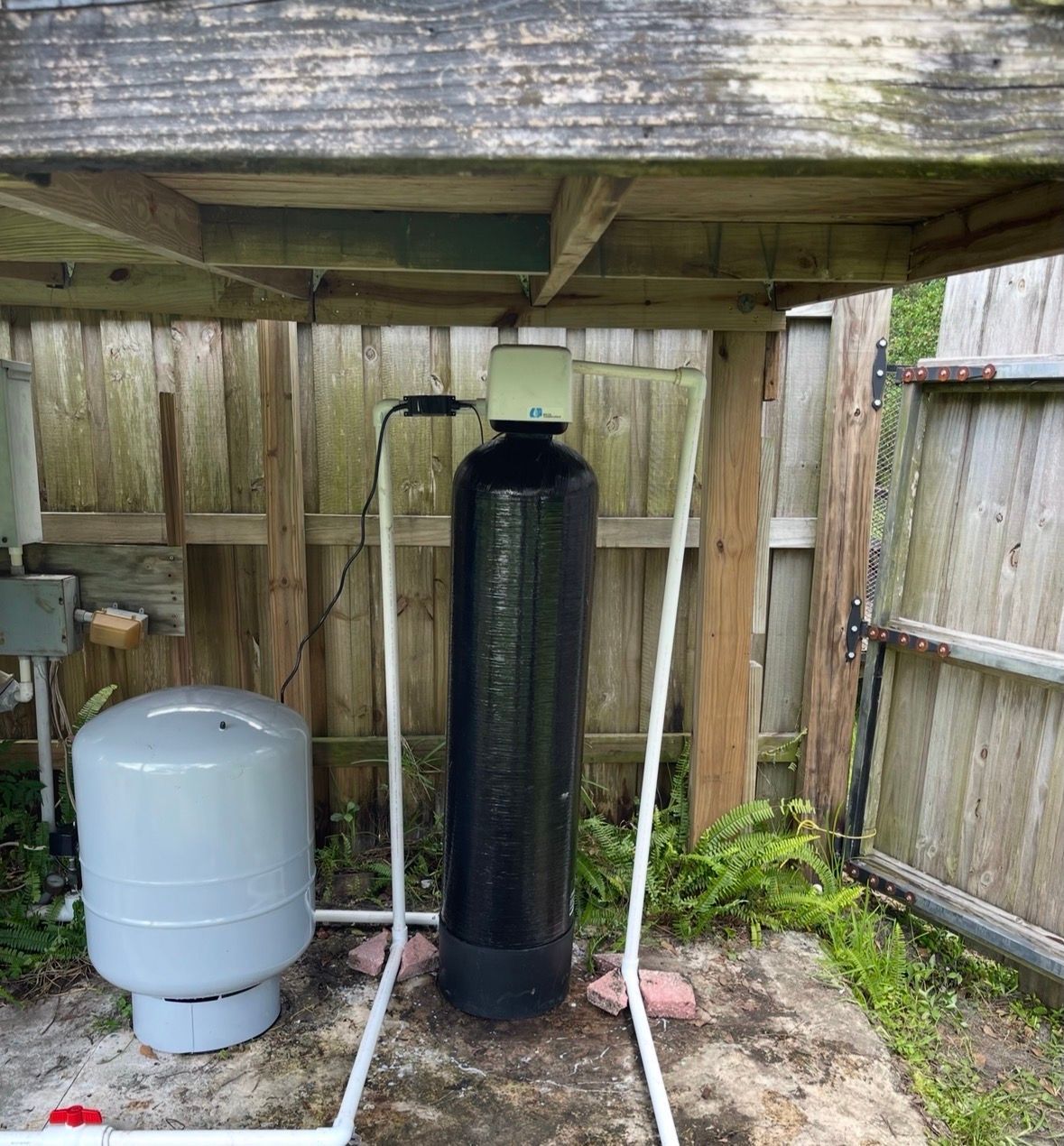 Covered outdoor utility area with a black water softener and gray tank beside a wooden fence