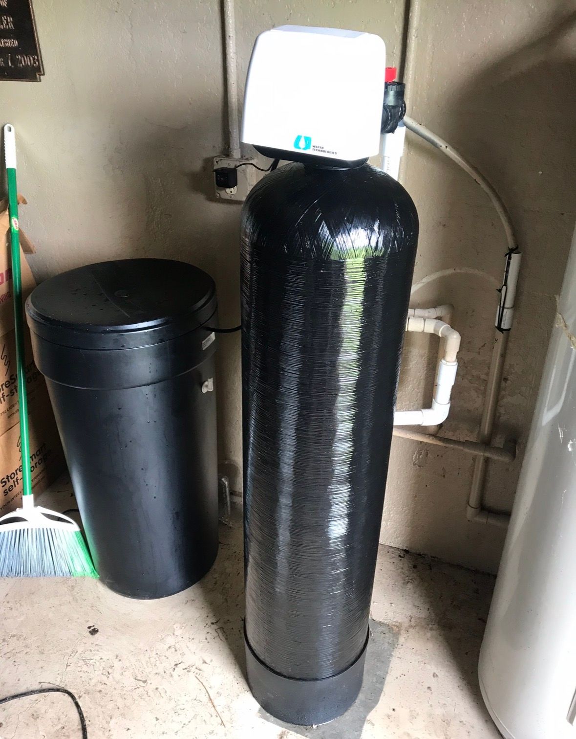 Black water softener tank with control head in a utility room, beside a black trash can.