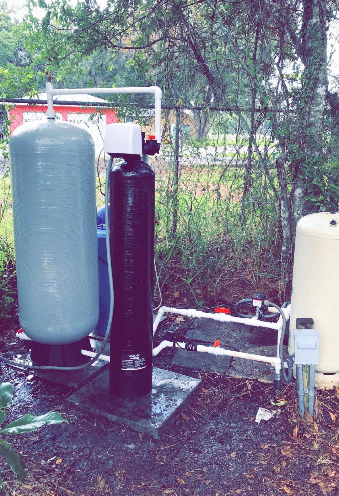 Outdoor water treatment equipment with two large tanks and pipes beside a fence and trees.