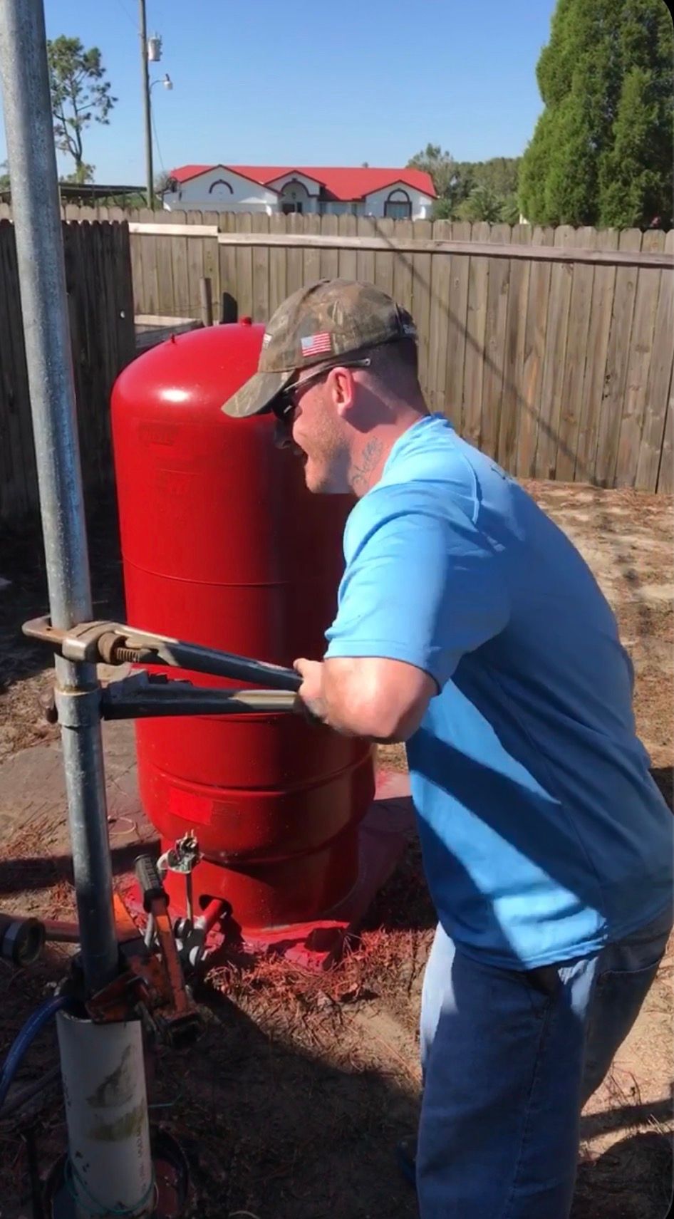 Worker in blue shirt adjusts red industrial tank beside a fenced yard.