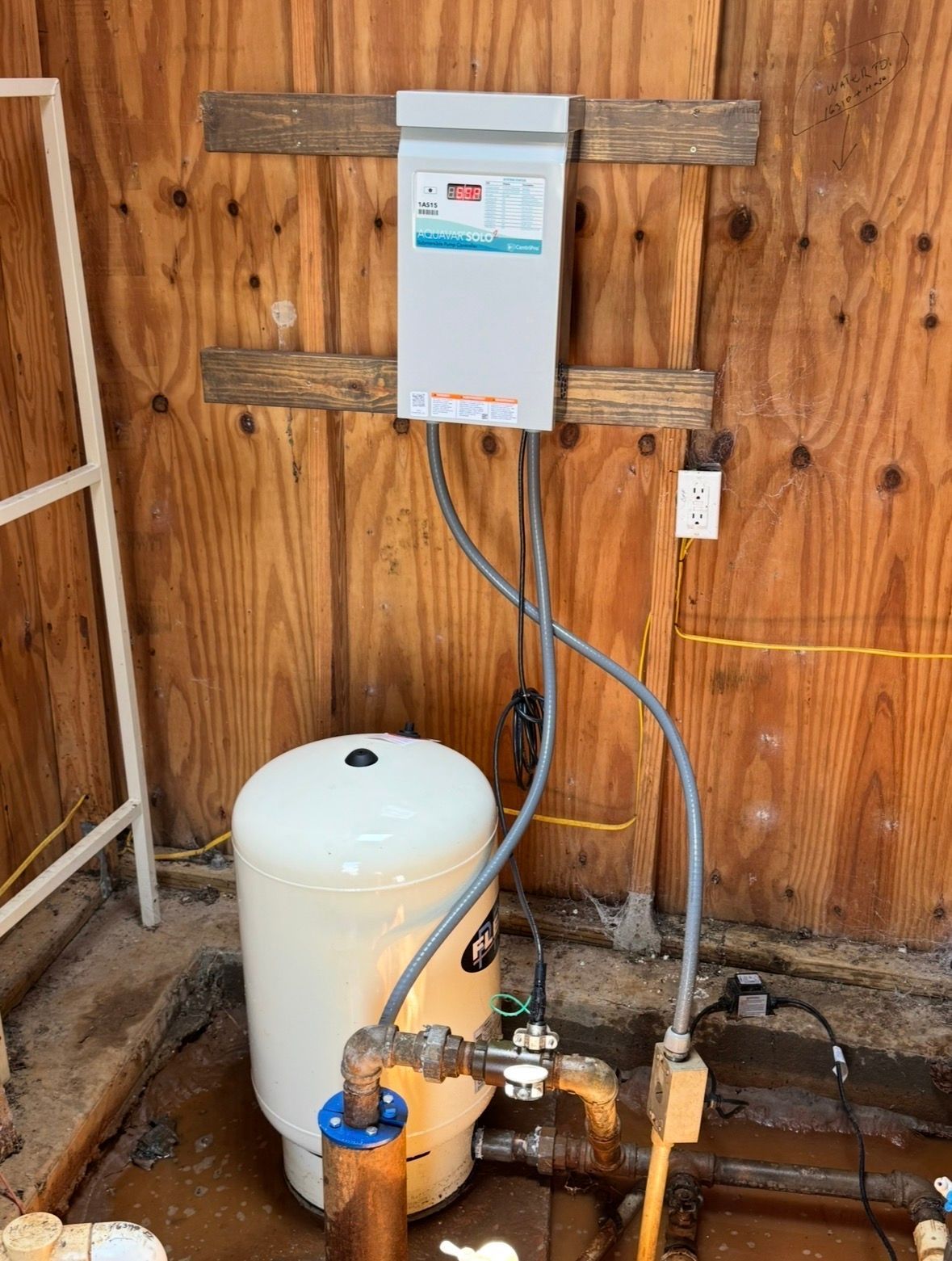 Water heater, pressure tank, and plumbing mounted on a wooden wall in a utility area.