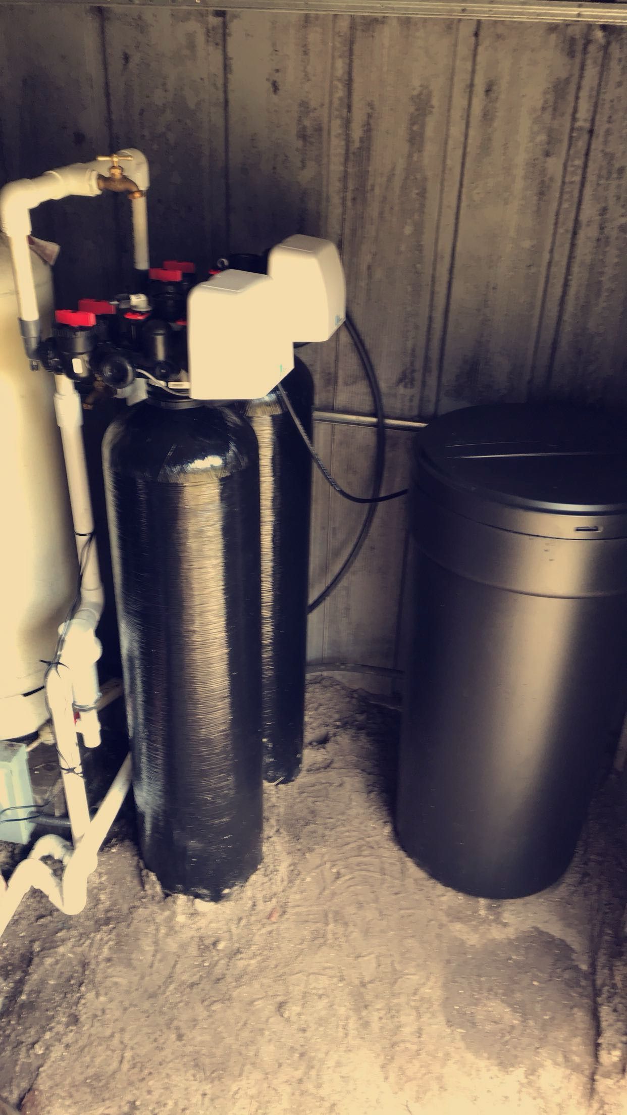 Water filtration tanks and plumbing equipment in a utility room, with two black cylindrical tanks against a concrete wall.