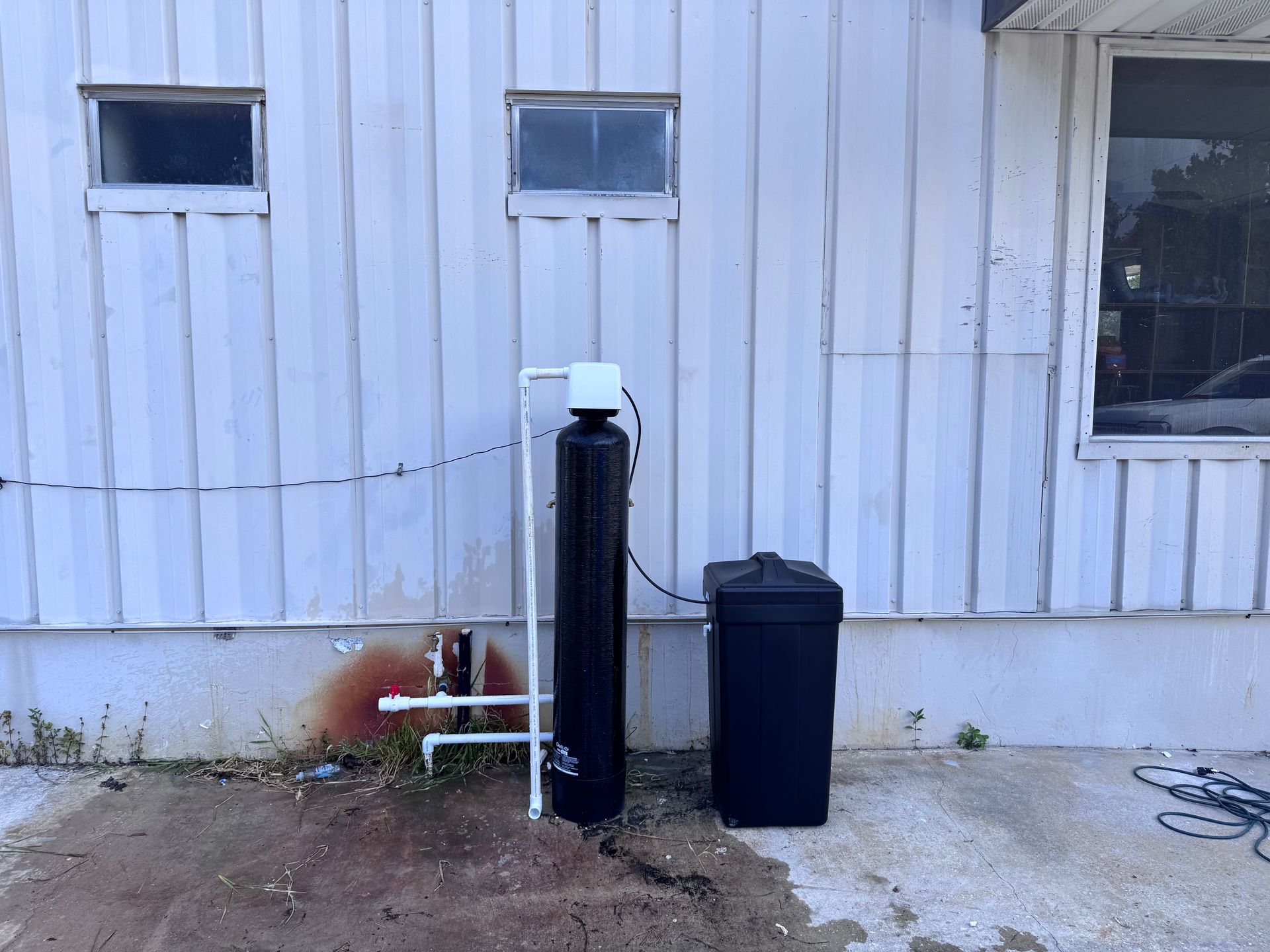 Outdoor utility equipment beside a white building, with a black post, bin, pipes, and windows