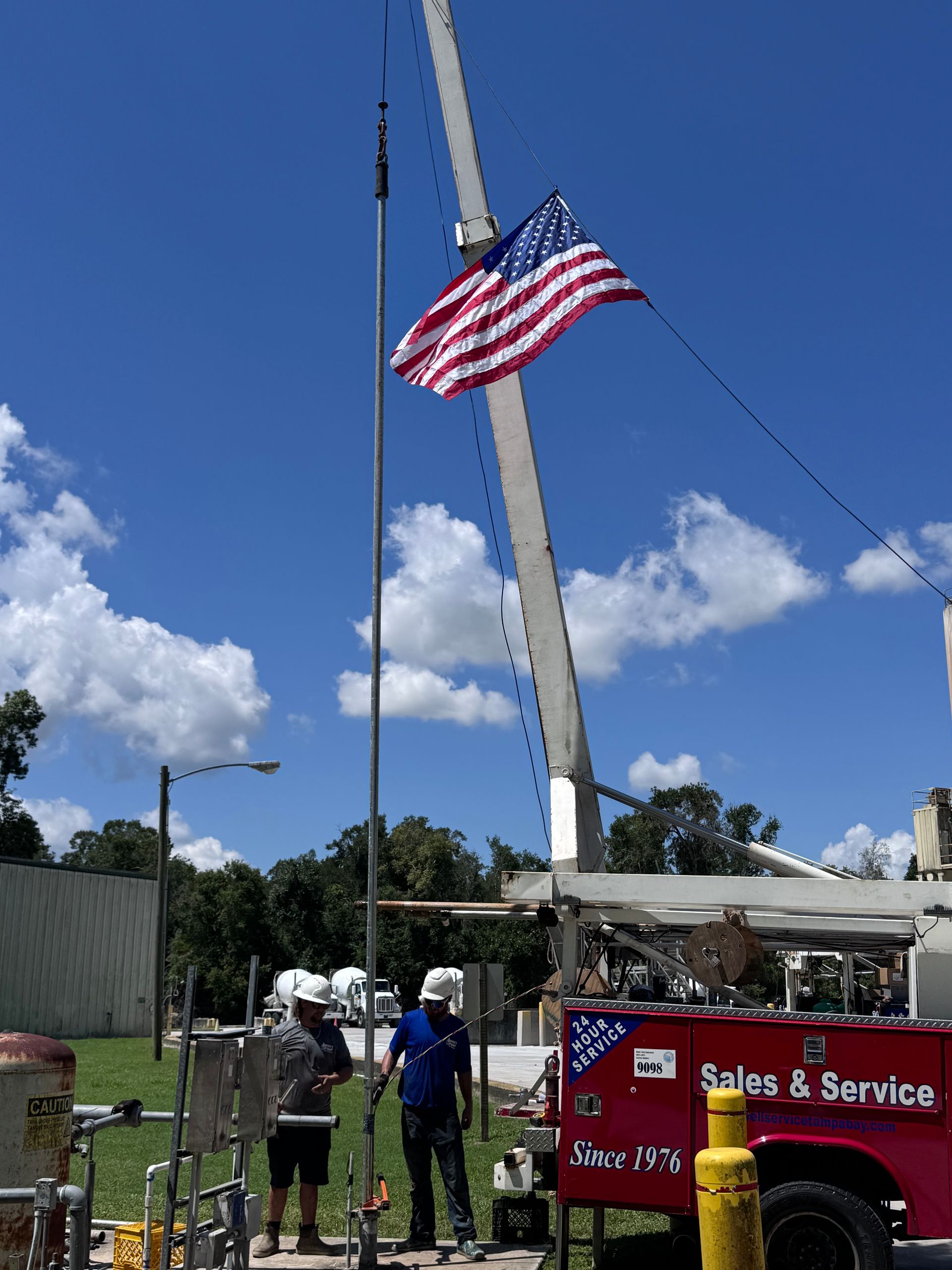 American flag waving on a tall crane under a blue sky, with workers and a service truck below.