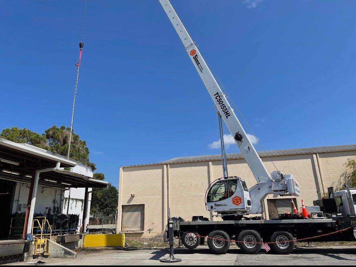 Mobile crane on a flatbed trailer beside a beige building under a clear blue sky.