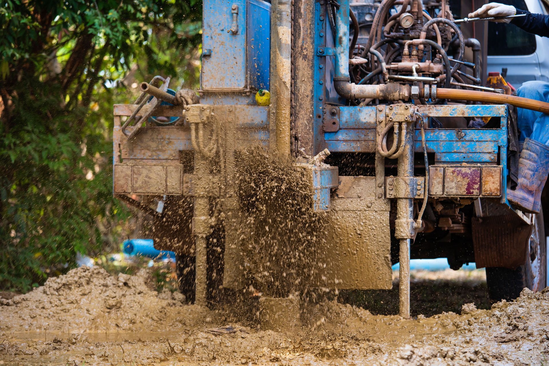 Blue drilling machine spraying mud and water on a dirt road beside trees