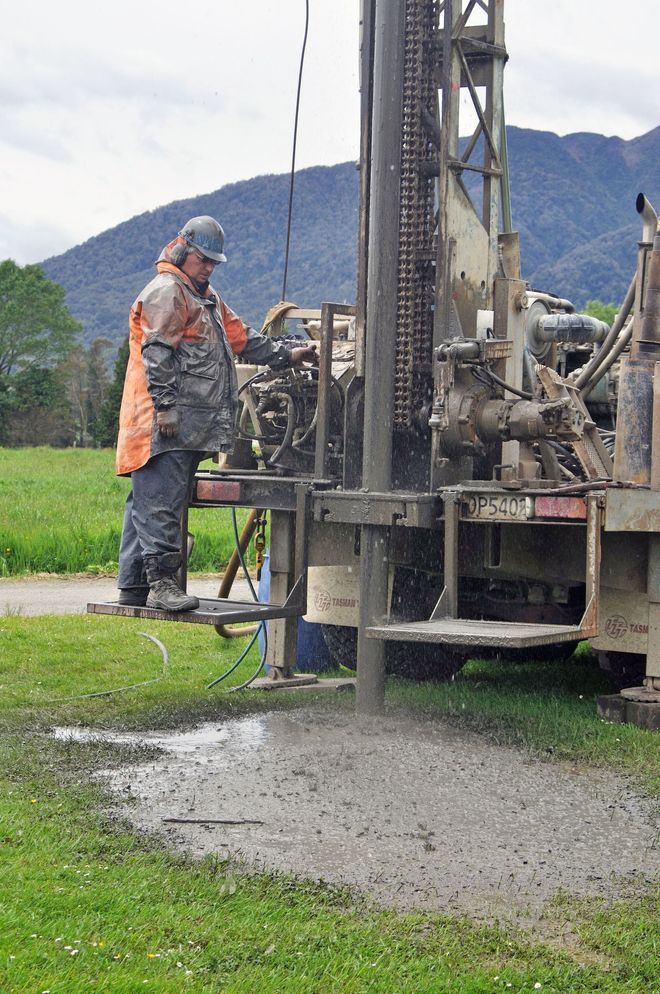 Worker in orange vest operating a drilling rig on a gravel site with mountains in the background