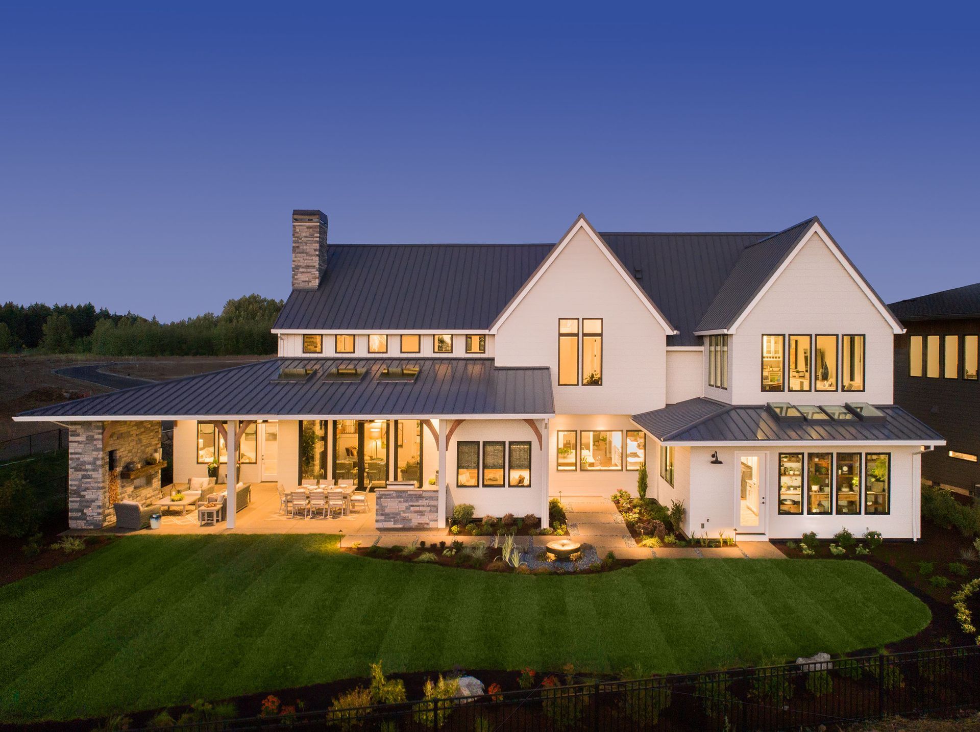 Large white house with lit windows and porch at dusk, set on a green lawn.