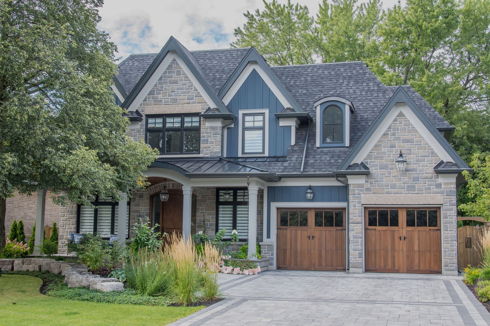Two-story stone house with dark roof, wood garage doors, and landscaped front yard.
