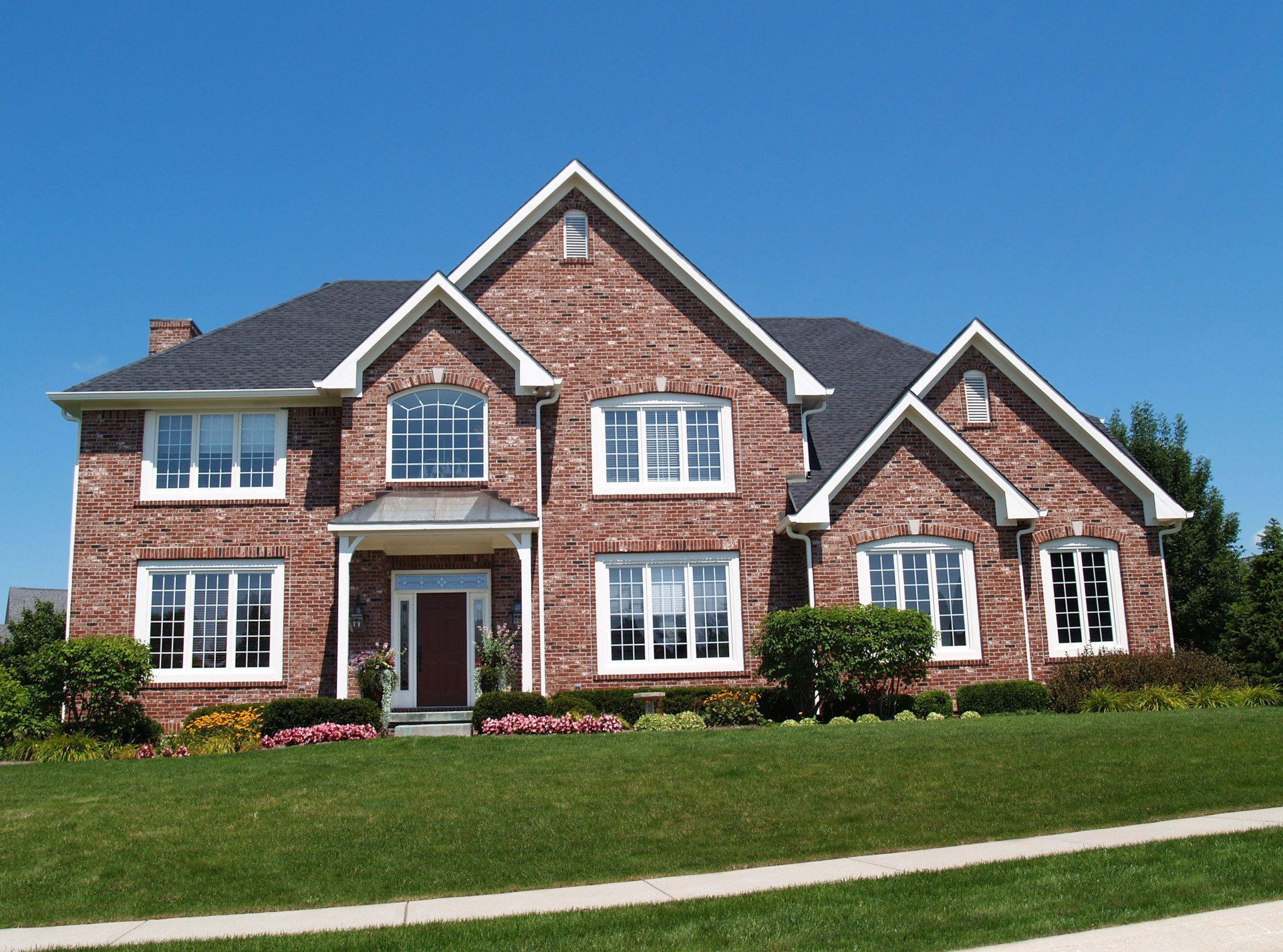 Large brick suburban house with white trim and manicured lawn under a blue sky.