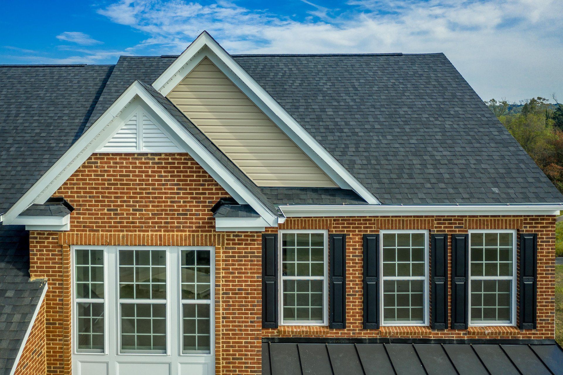 Brick house facade with gray shingled roof, white trim, and front windows under a blue sky.