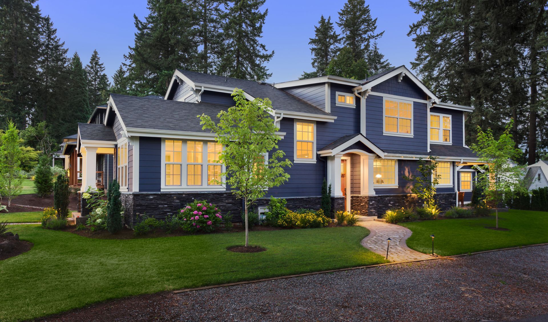 Blue-gray two-story house with lit windows and a front porch, surrounded by trees and a green lawn at dusk.
