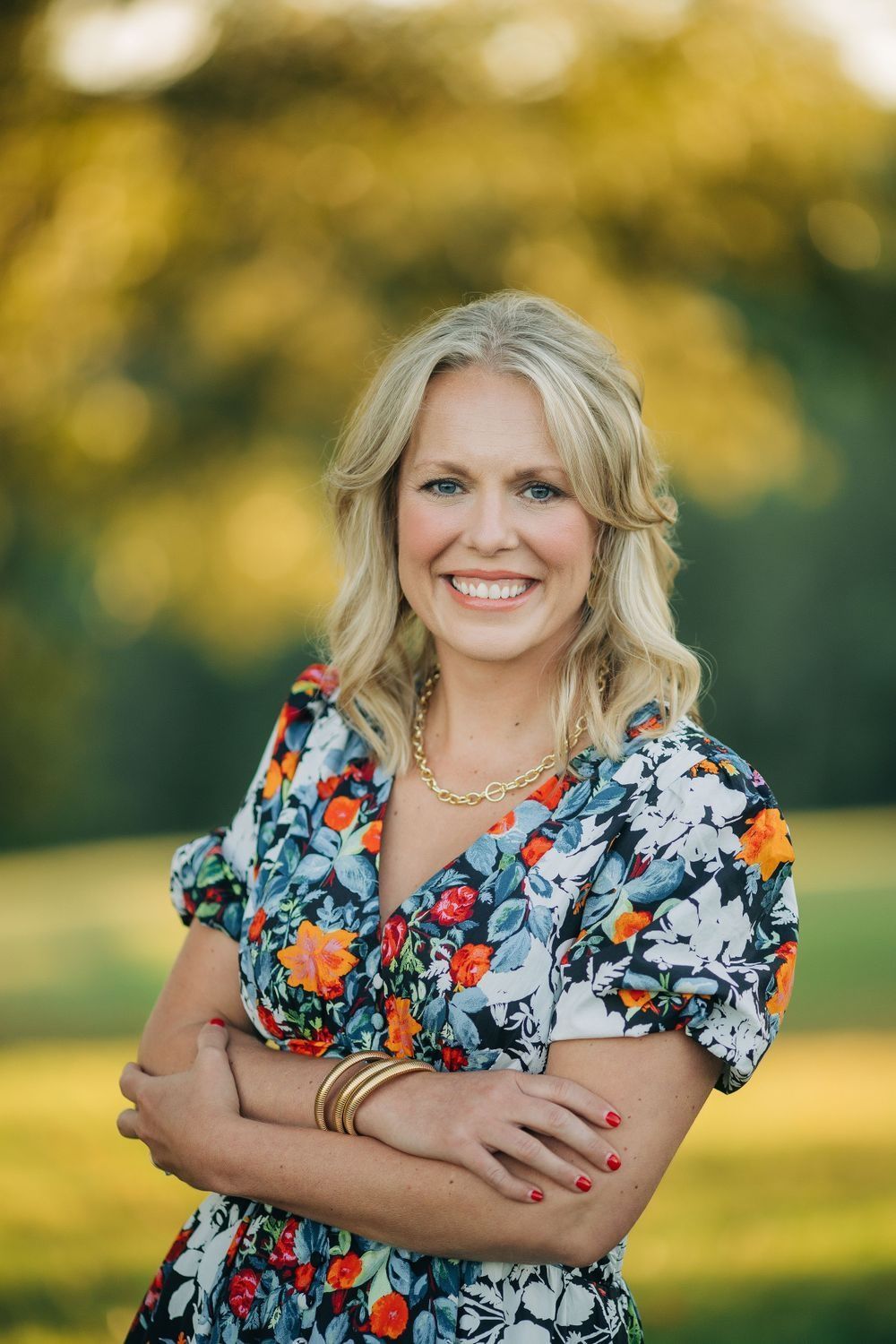 Smiling person with crossed arms wearing a floral shirt outdoors with blurred trees in the background