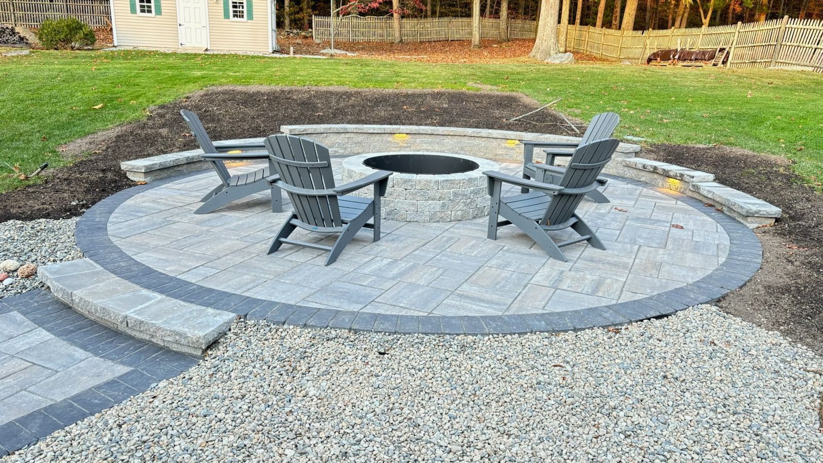Outdoor stone fire pit seating area with four gray chairs on a gravel patio in a yard