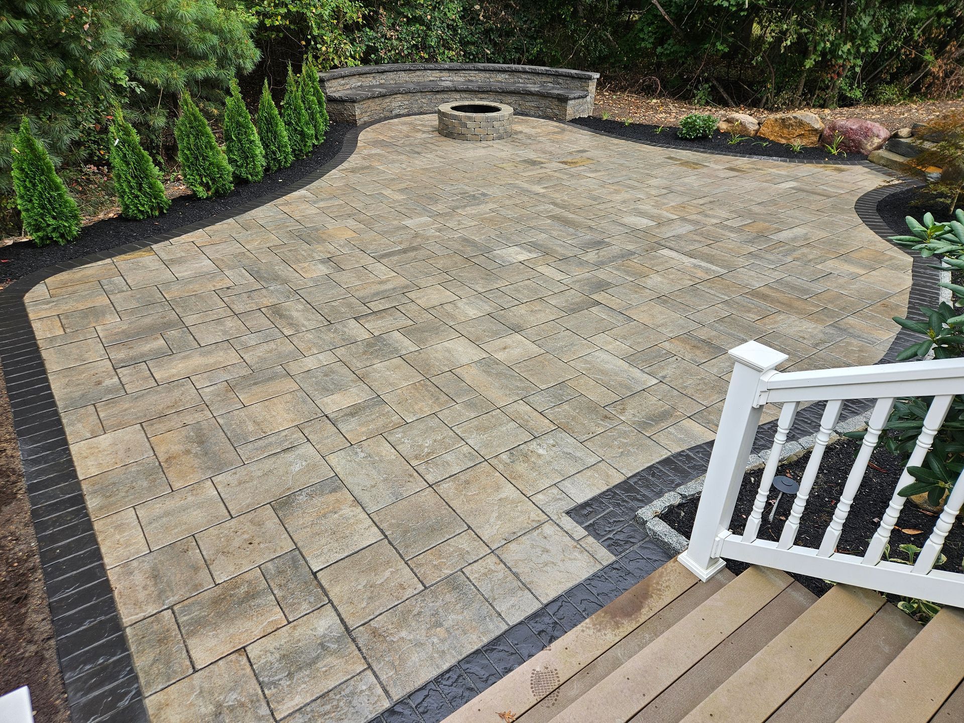 Curved brick patio with built-in fire pit, bordered by landscaping and white deck railing