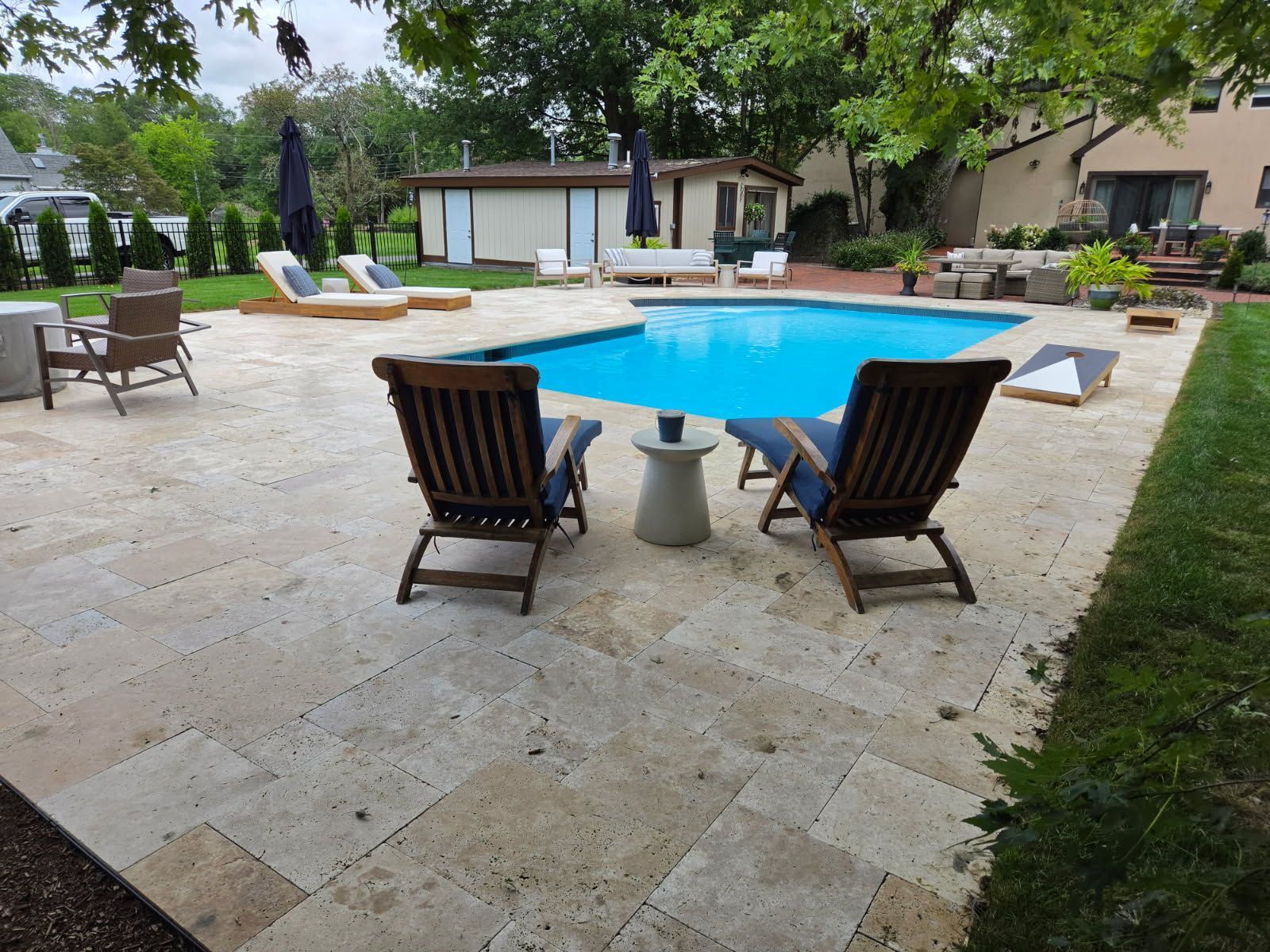 Backyard patio with a blue swimming pool, lounge chairs, and a small table under trees.