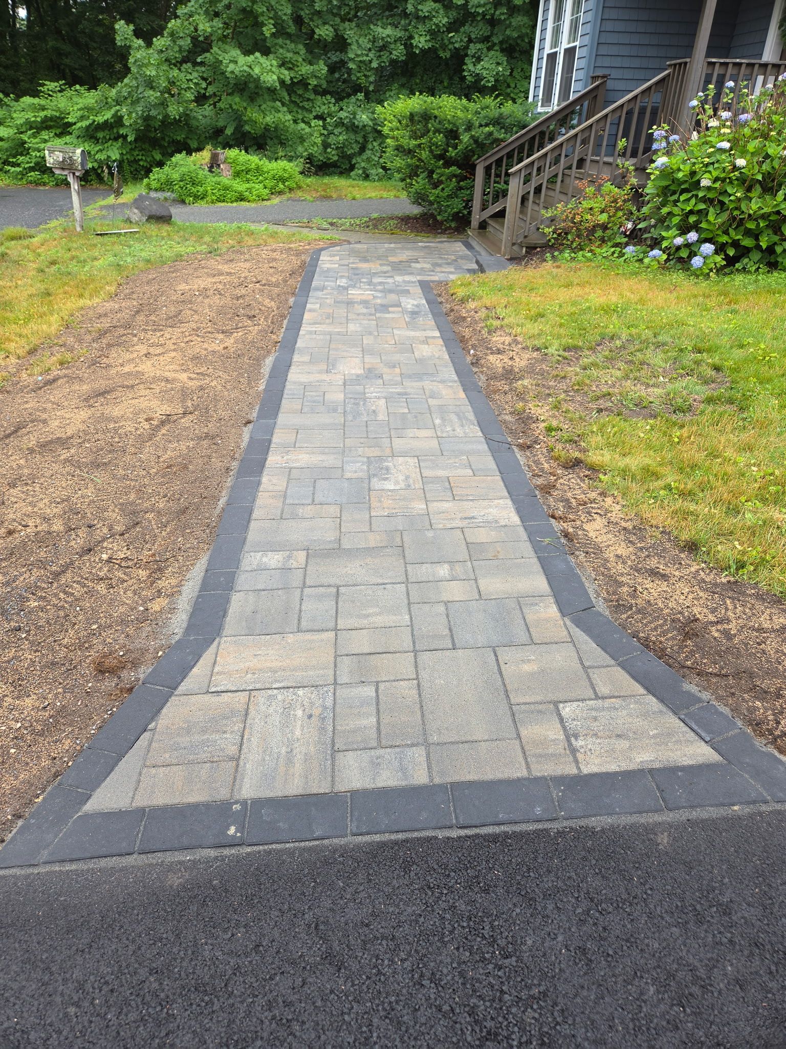 Stone walkway leading from a road to a house, bordered by mulch and green shrubs.