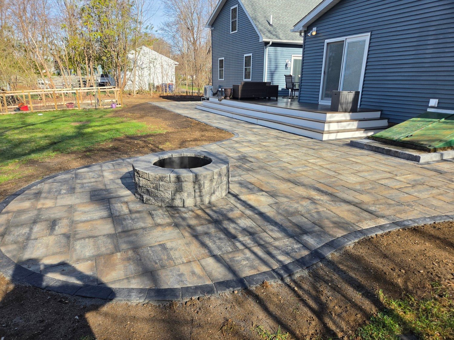 A stone fire pit sits on a paved patio adjacent to a blue house with white steps.