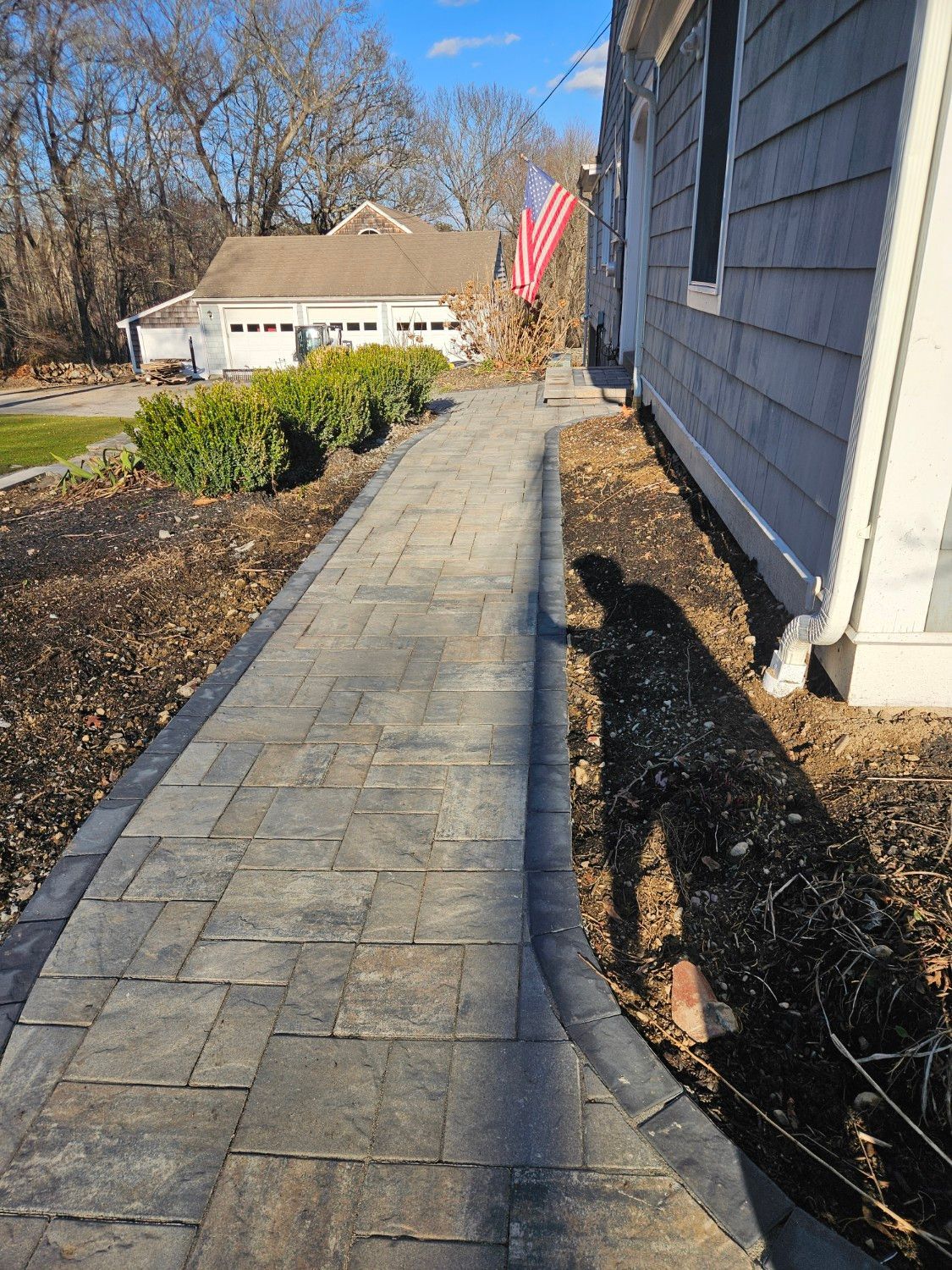 A paved walkway leads alongside a house with gray siding toward a detached garage, with landscaping shrubs to the left.
