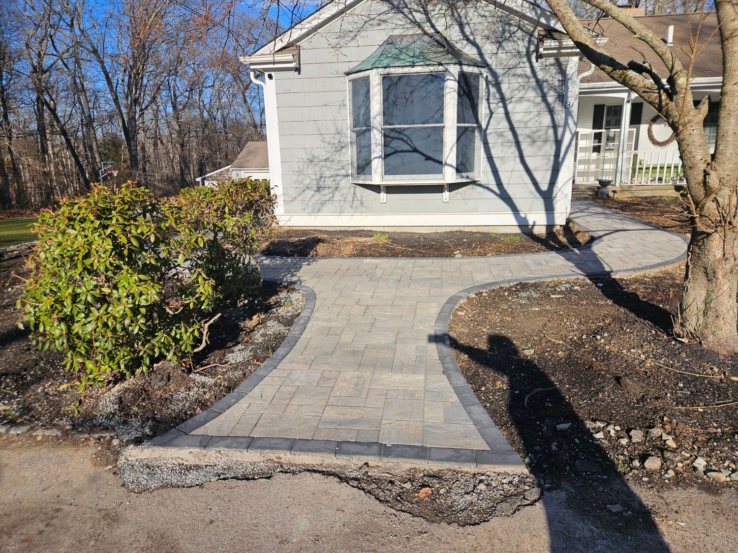 A paved walkway with a dark border leads from an asphalt driveway to the front of a light gray house with a bay window.