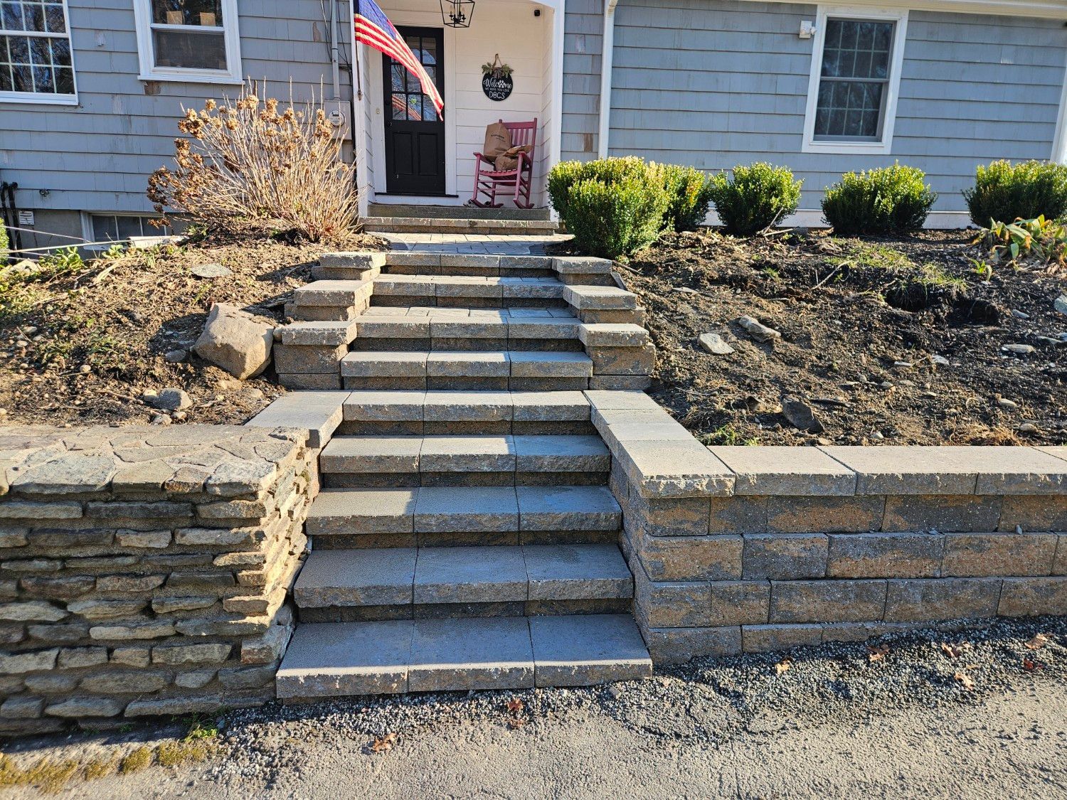 Gray stone steps lead up a terraced garden to the entrance of a house with light gray siding and an American flag.