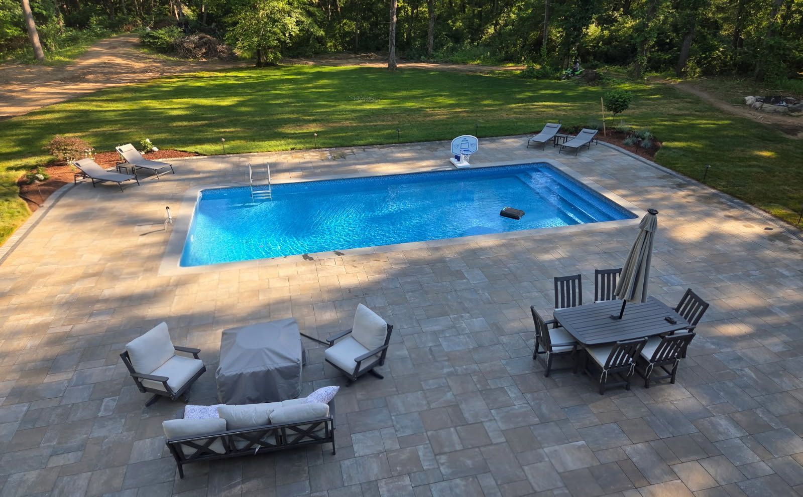 Backyard patio with a rectangular in-ground pool, lounge chairs, and a dining table surrounded by trees