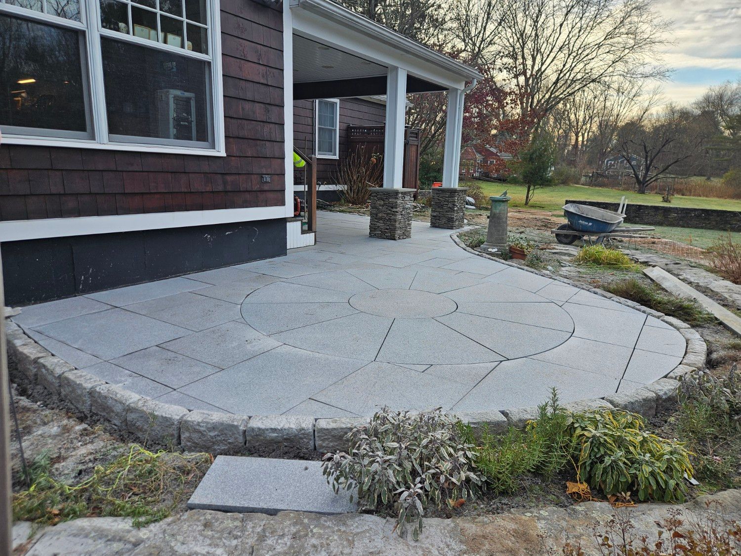 Stone patio with curved edge, by a house. Ground-level view. Brown siding, white columns, and plants.