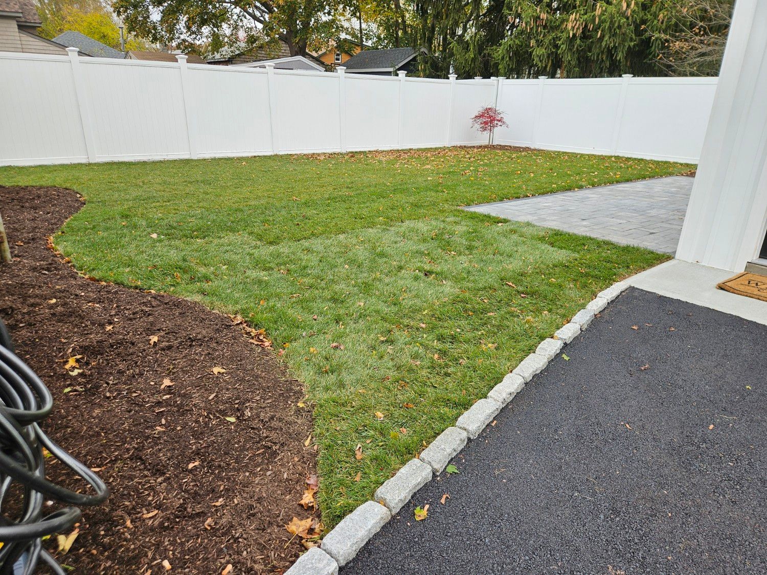 A backyard with a white fence, green grass, and a mulch bed. A paved driveway borders the lawn.