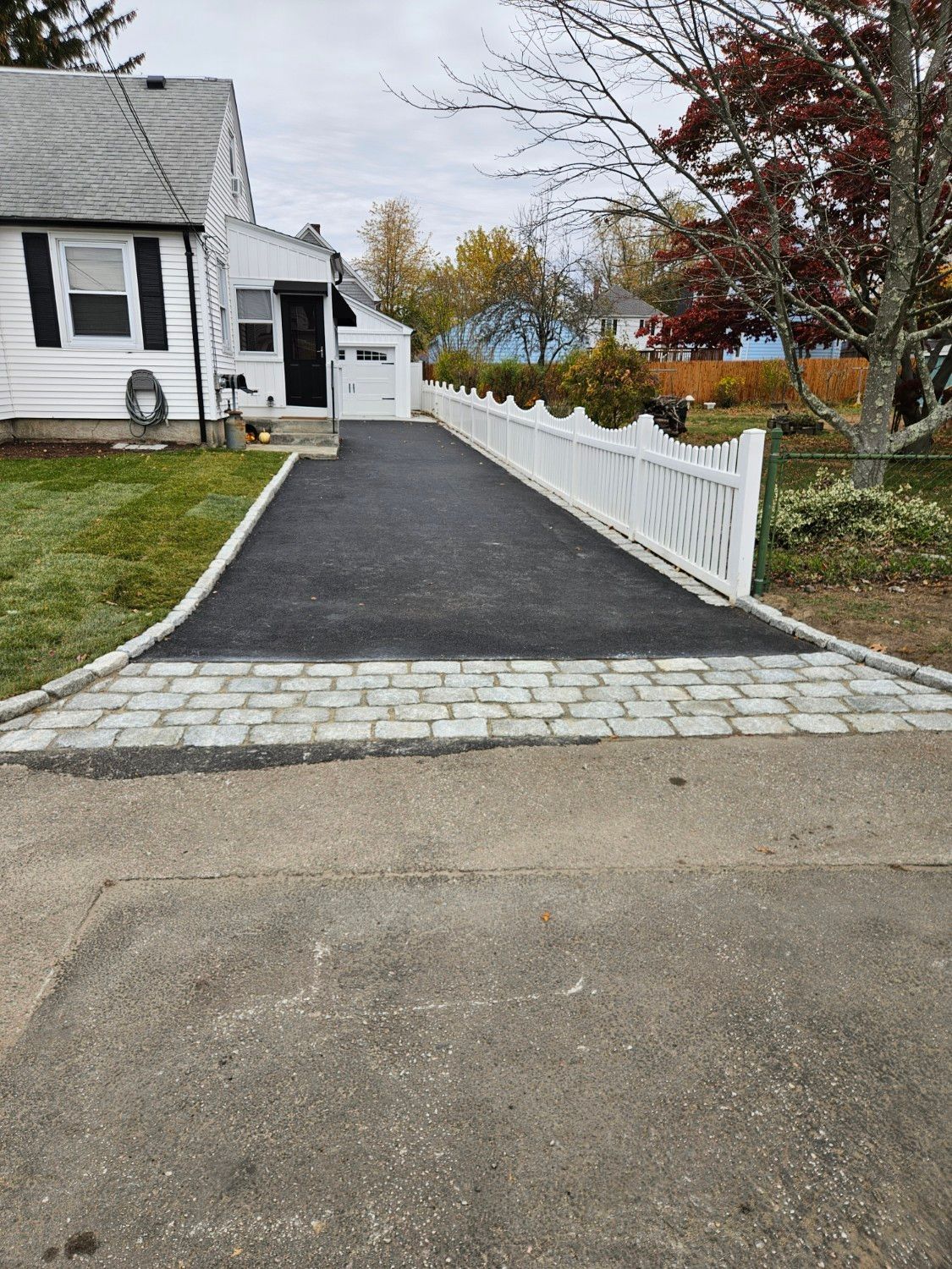 Asphalt driveway bordered by a white fence and a gray stone section, leading to a white house with a black front door.