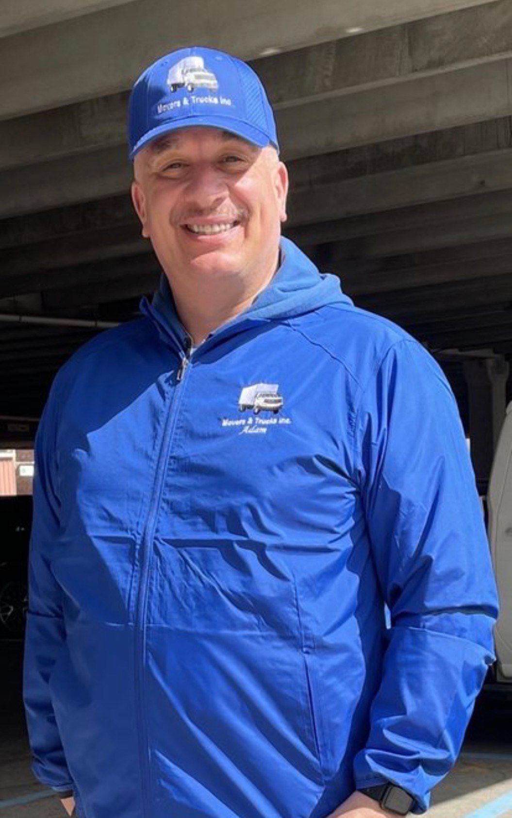 Man in blue cap and jacket, smiling. Outside, possibly a parking garage.