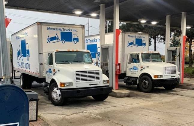 Two white moving trucks parked under a gas station canopy. Trucks have