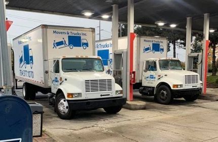 Two white moving trucks parked under a gas station canopy. Trucks have 
