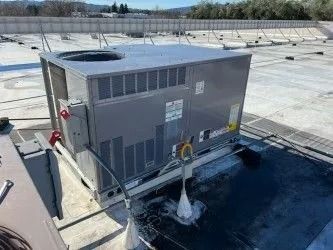 A grey rooftop HVAC unit sits on a flat, white roof surface under a clear blue sky.