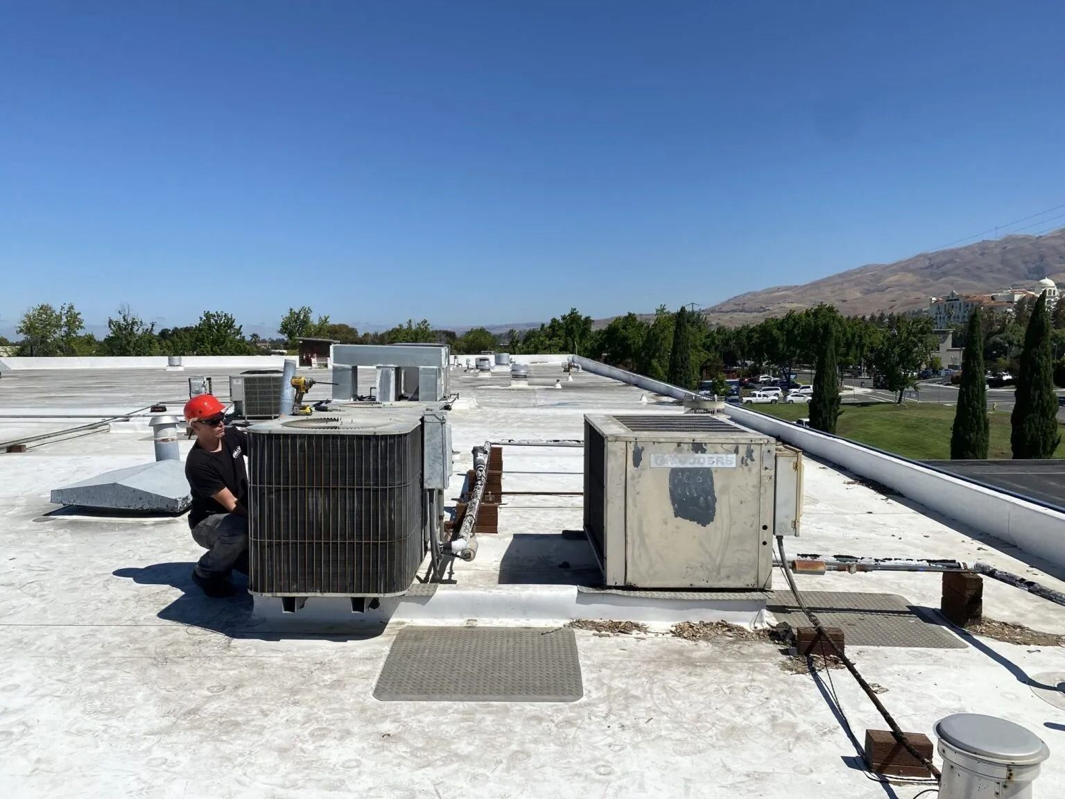 Technician in a red hard hat kneeling to service HVAC equipment on a flat building roof under a bright blue sky.